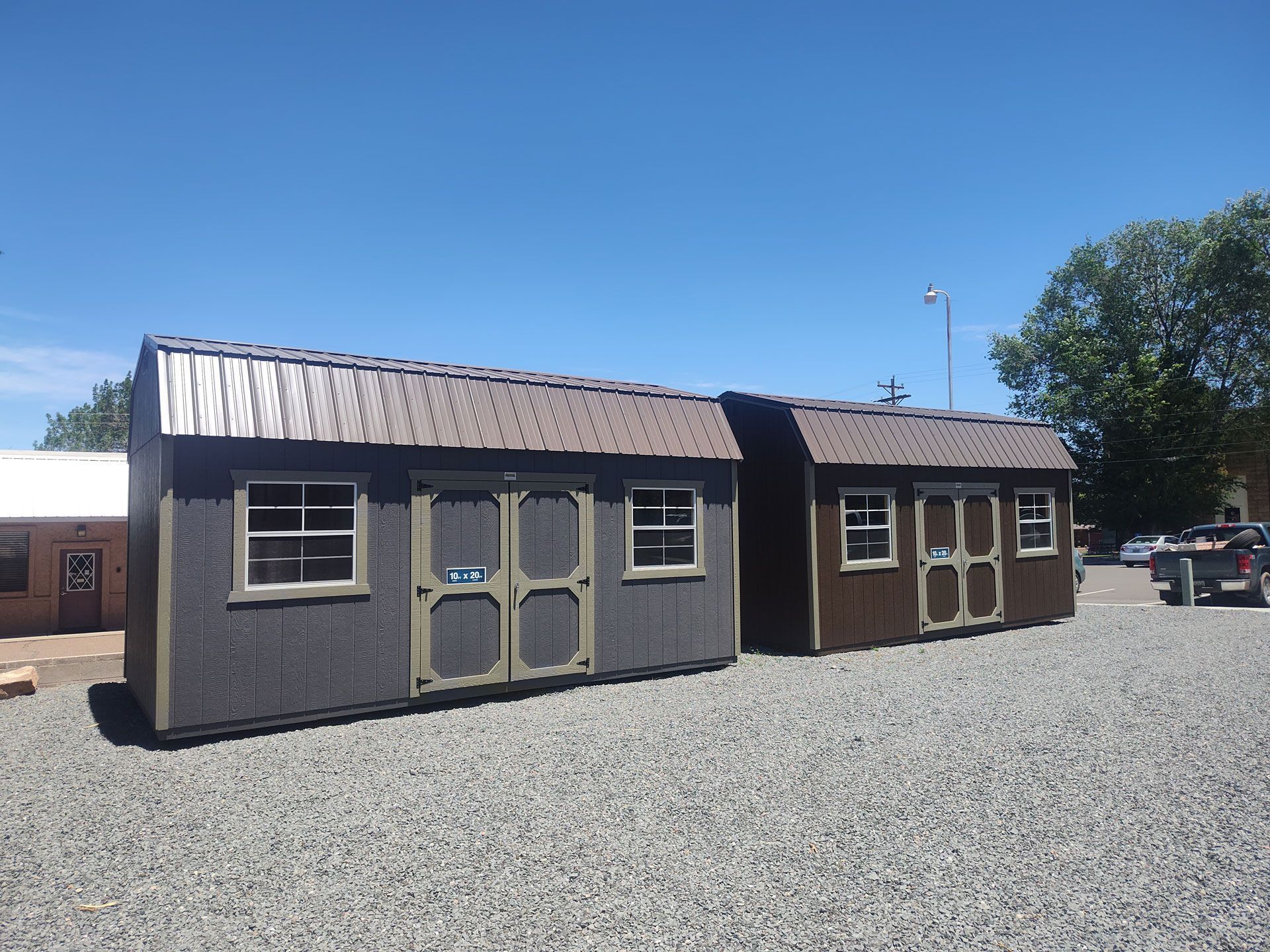 Two small gray sheds with brown roofs on a gravel lot under a clear blue sky