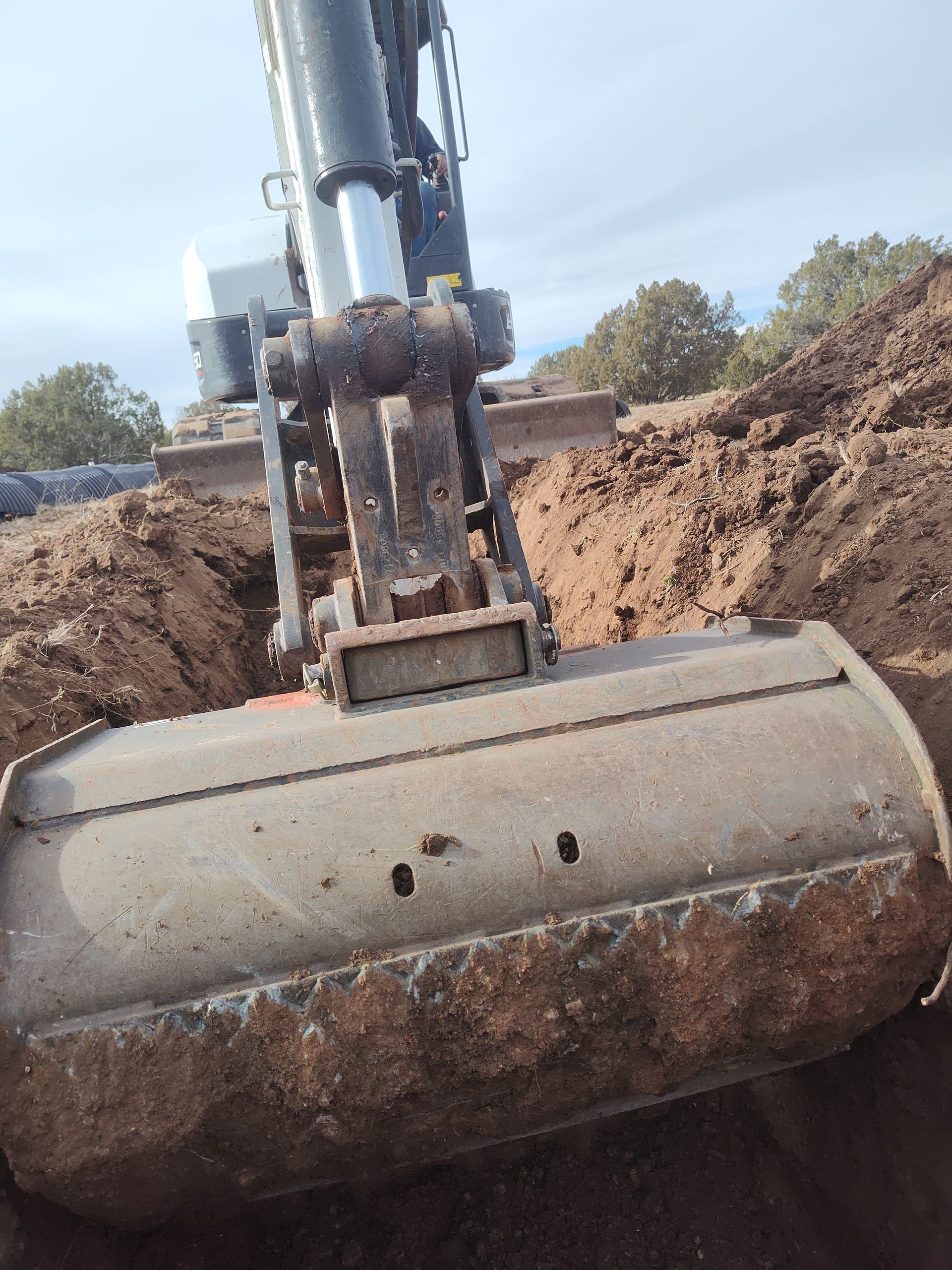Excavator bucket lifting a large dirt-covered metal tank at a construction site.