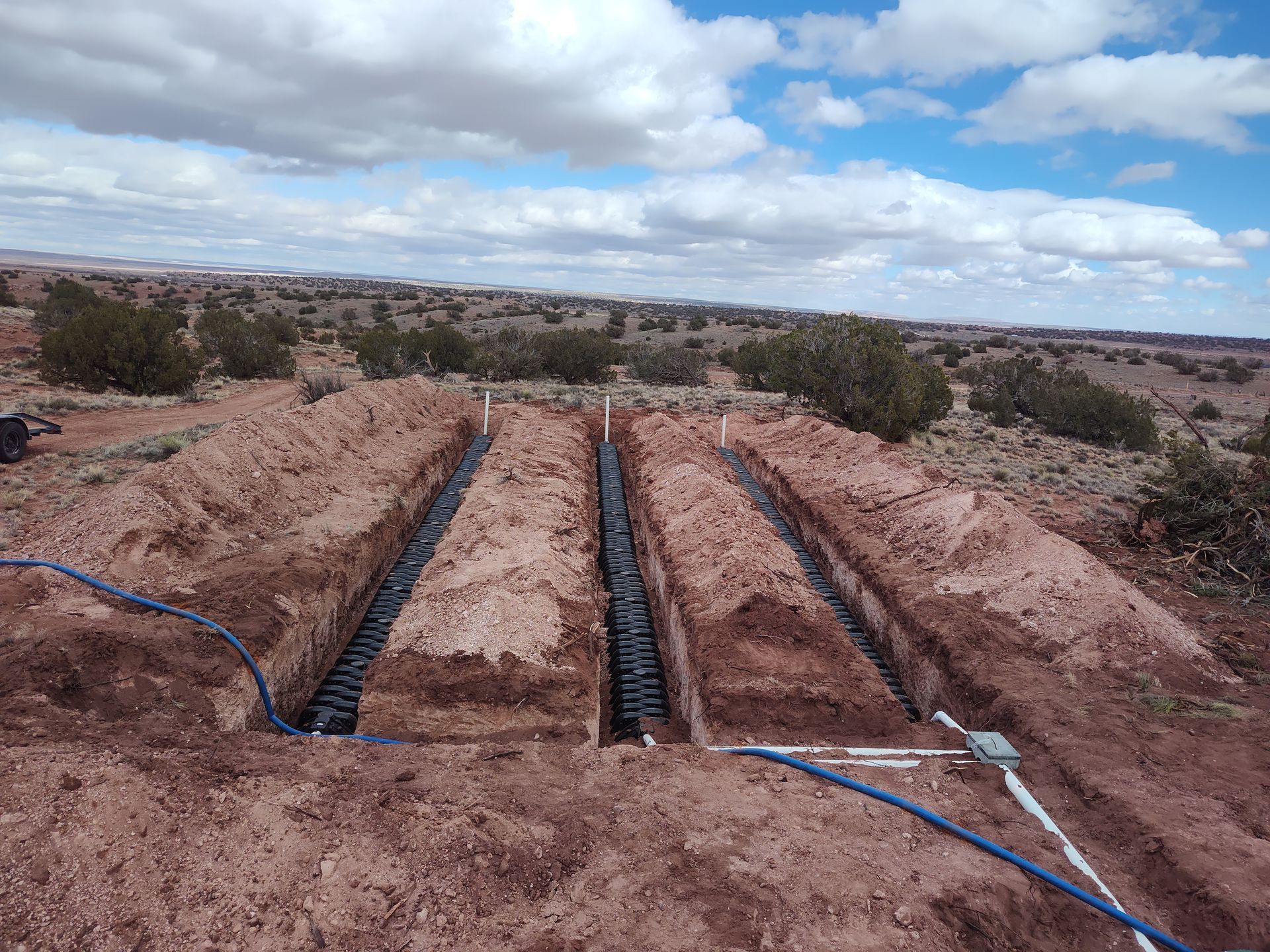 Trenches with black pipes in a red dirt landscape under a cloudy sky