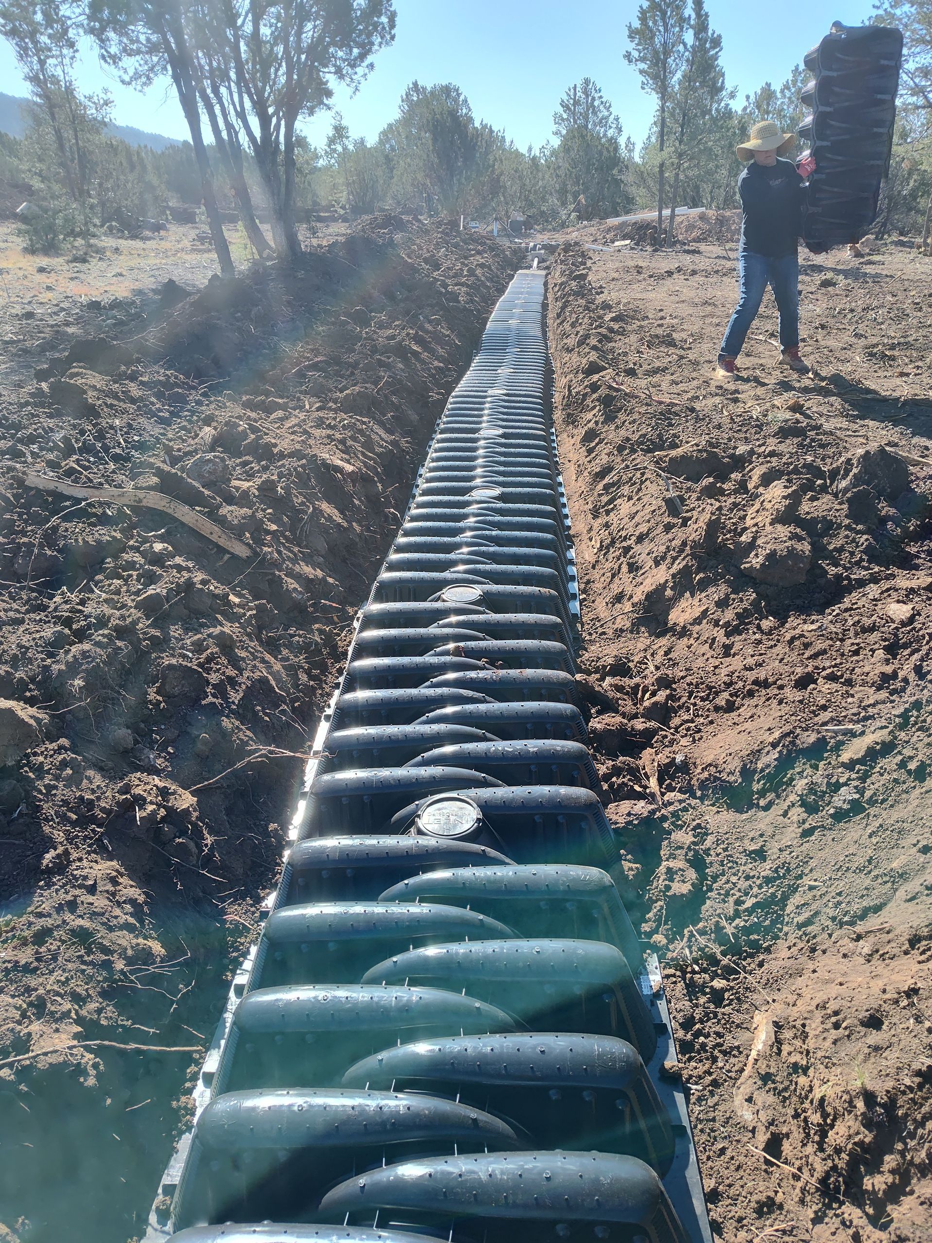 A large black pipeline running through a dirt trench beside a person standing on the bank.