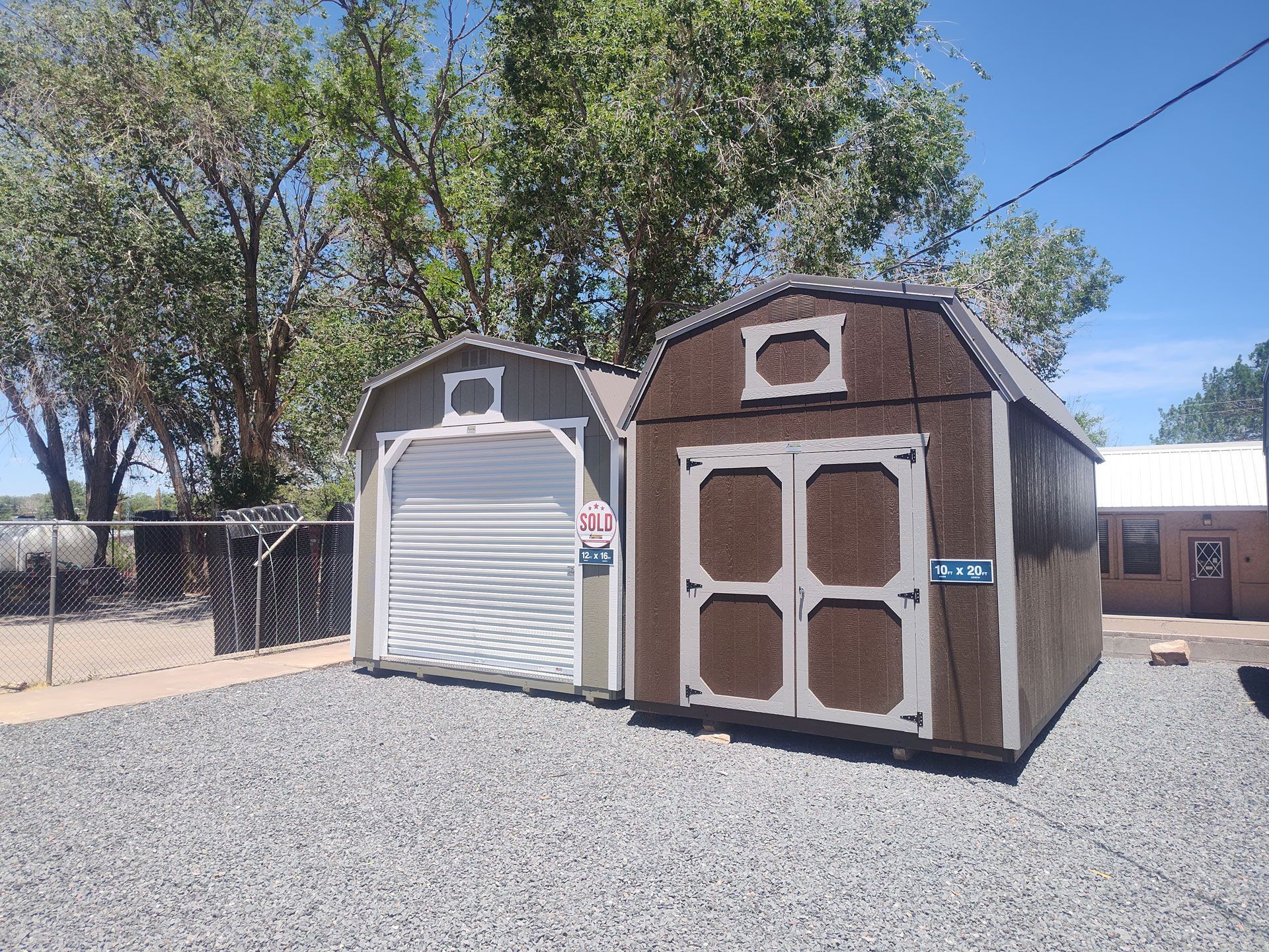 Two small storage sheds on a gravel lot, one gray and one brown, with trees and a clear sky behind them.