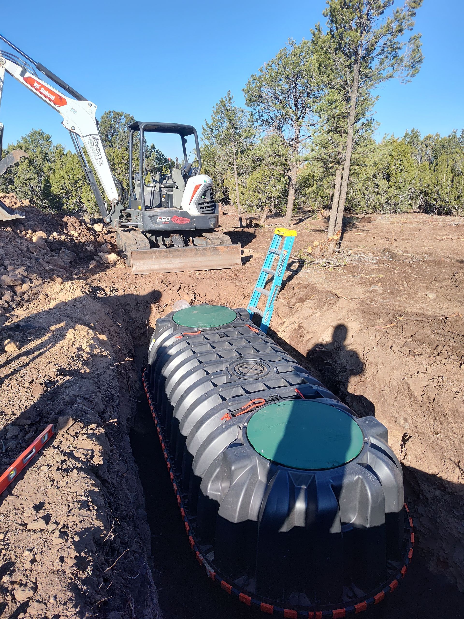 Gray underground tank being lowered into a trench at a construction site, with excavator and worker nearby.