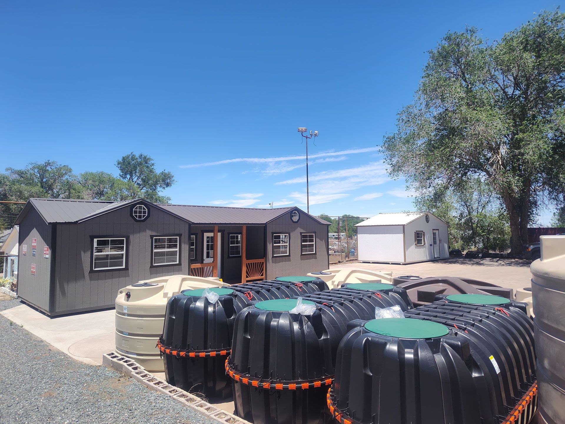 Gray modular buildings and large black water tanks in a sunny gravel yard under a blue sky
