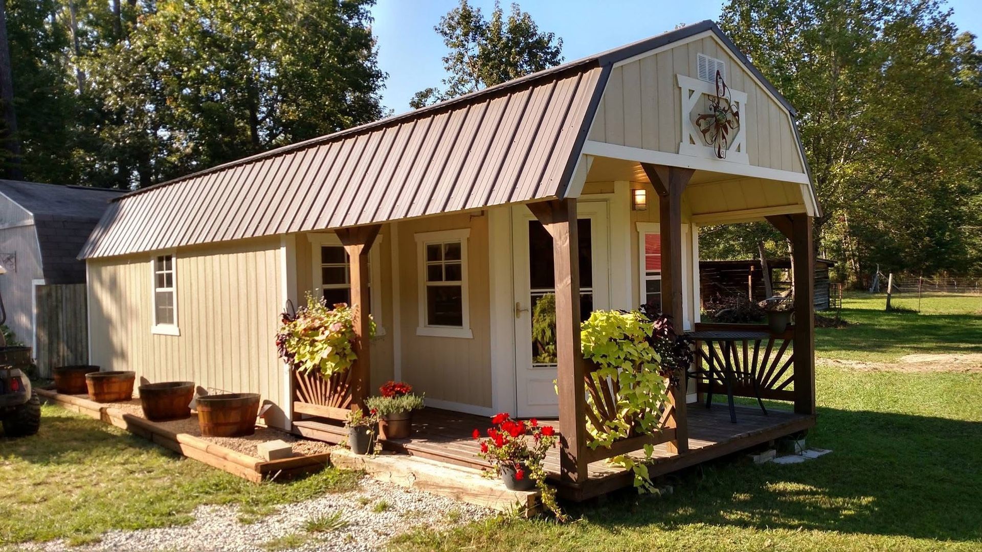 Small beige cabin with brown metal roof, porch, and potted plants in a sunny yard.