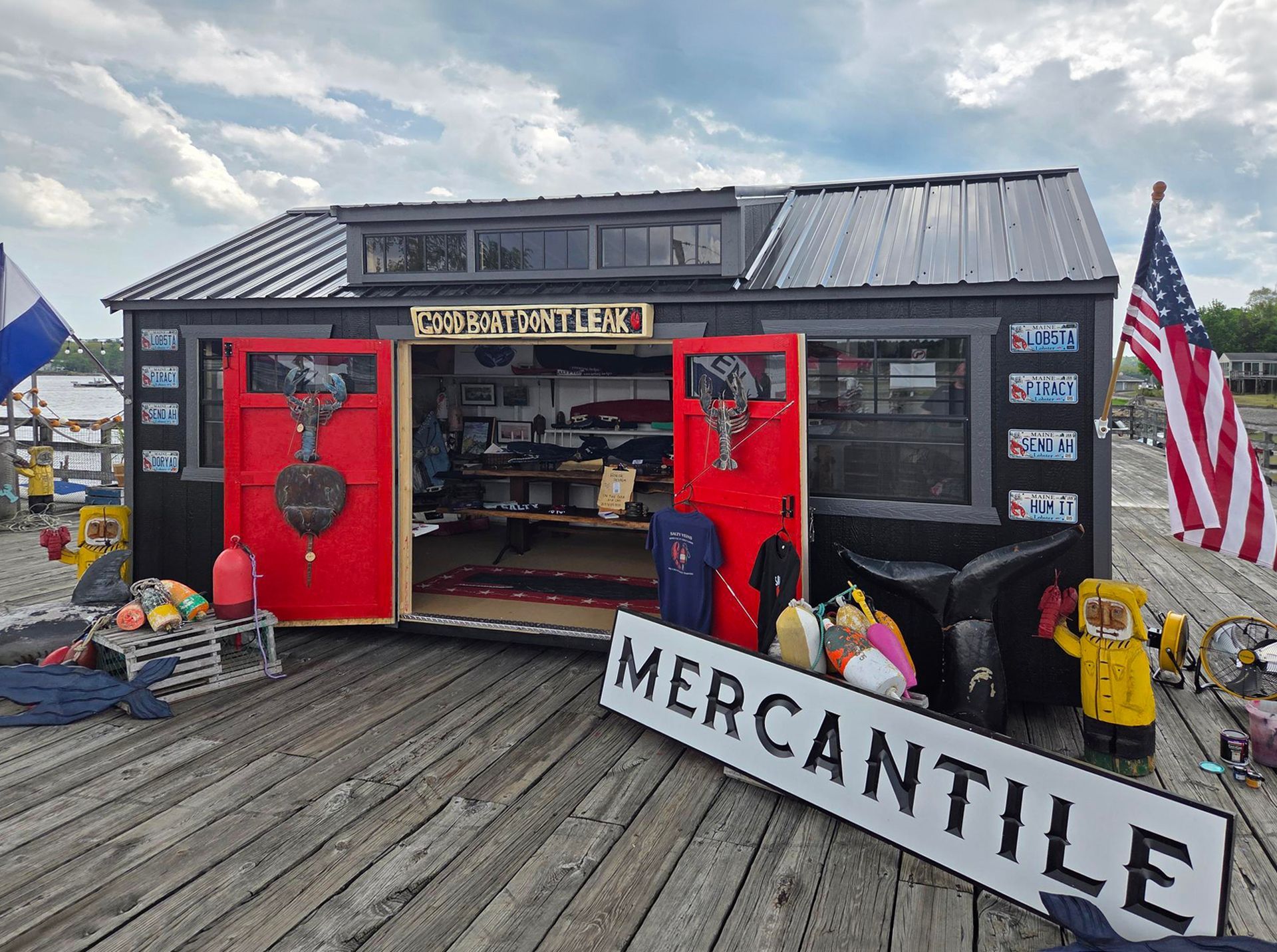 Black mercantile shack with open red doors, merchandise inside, and a large American flag on a wooden deck