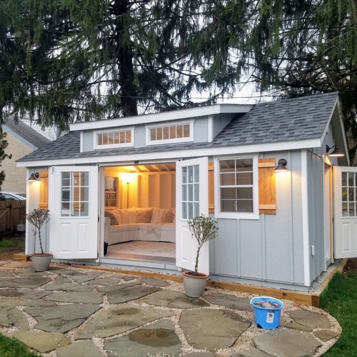 Small gray cottage with warm porch lights, white trim, stone patio, and potted plants at dusk