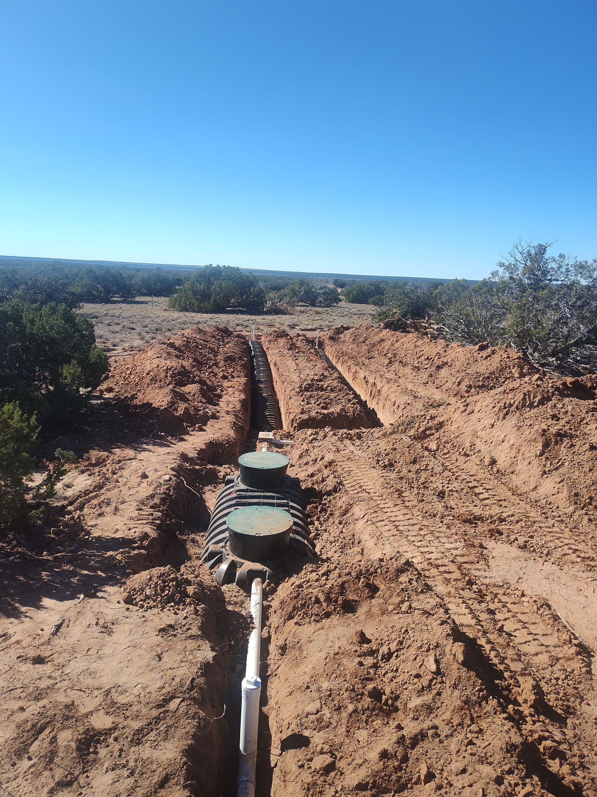 Trench in reddish dirt with exposed utility pipe and circular access covers under a clear blue sky