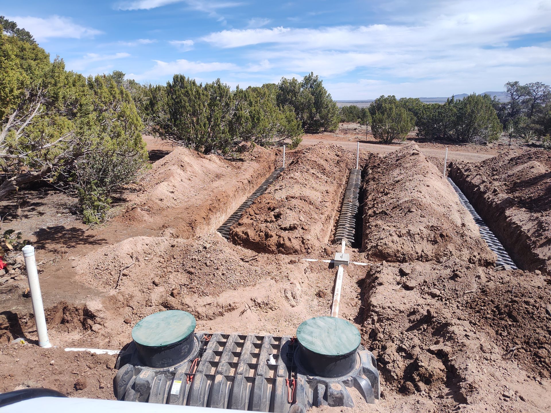 Rural dirt excavation with trenches, pipes, and green utility covers beside trees