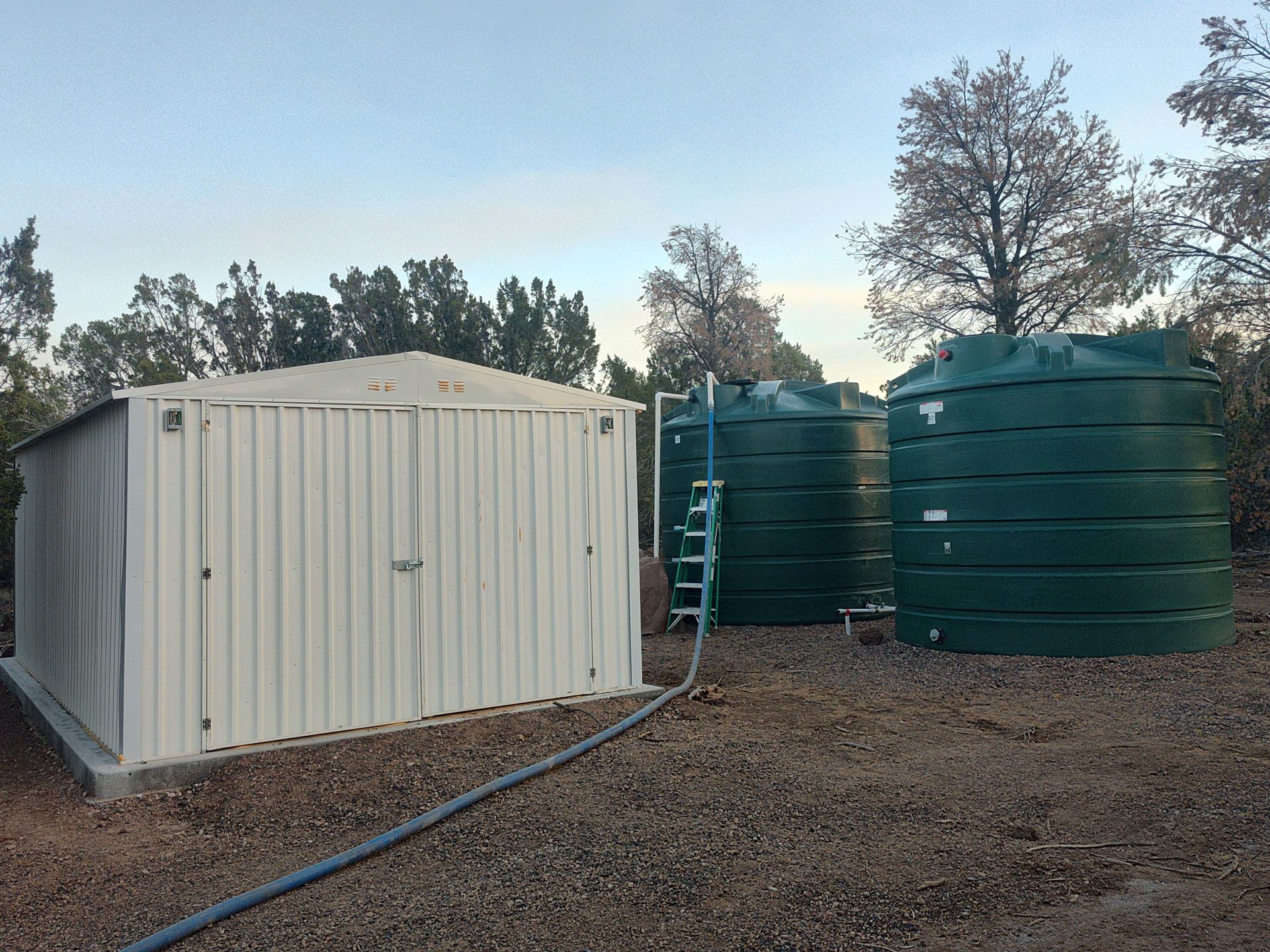 White shed beside large green water tanks in a gravel yard at dusk