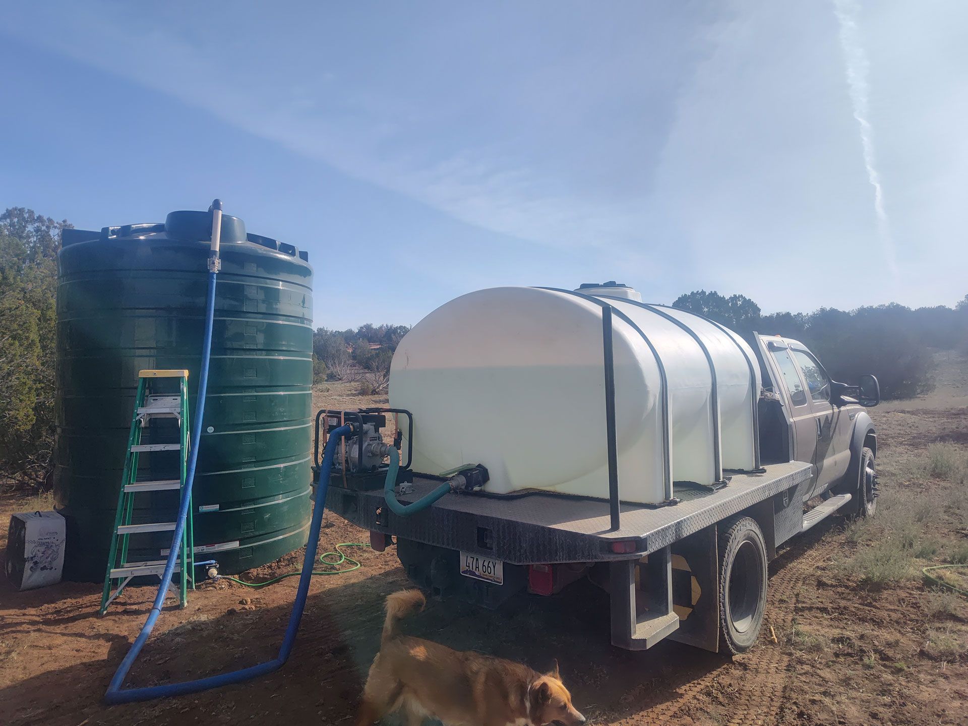 Water truck with white tank beside large green water tanks in a rural outdoor area.