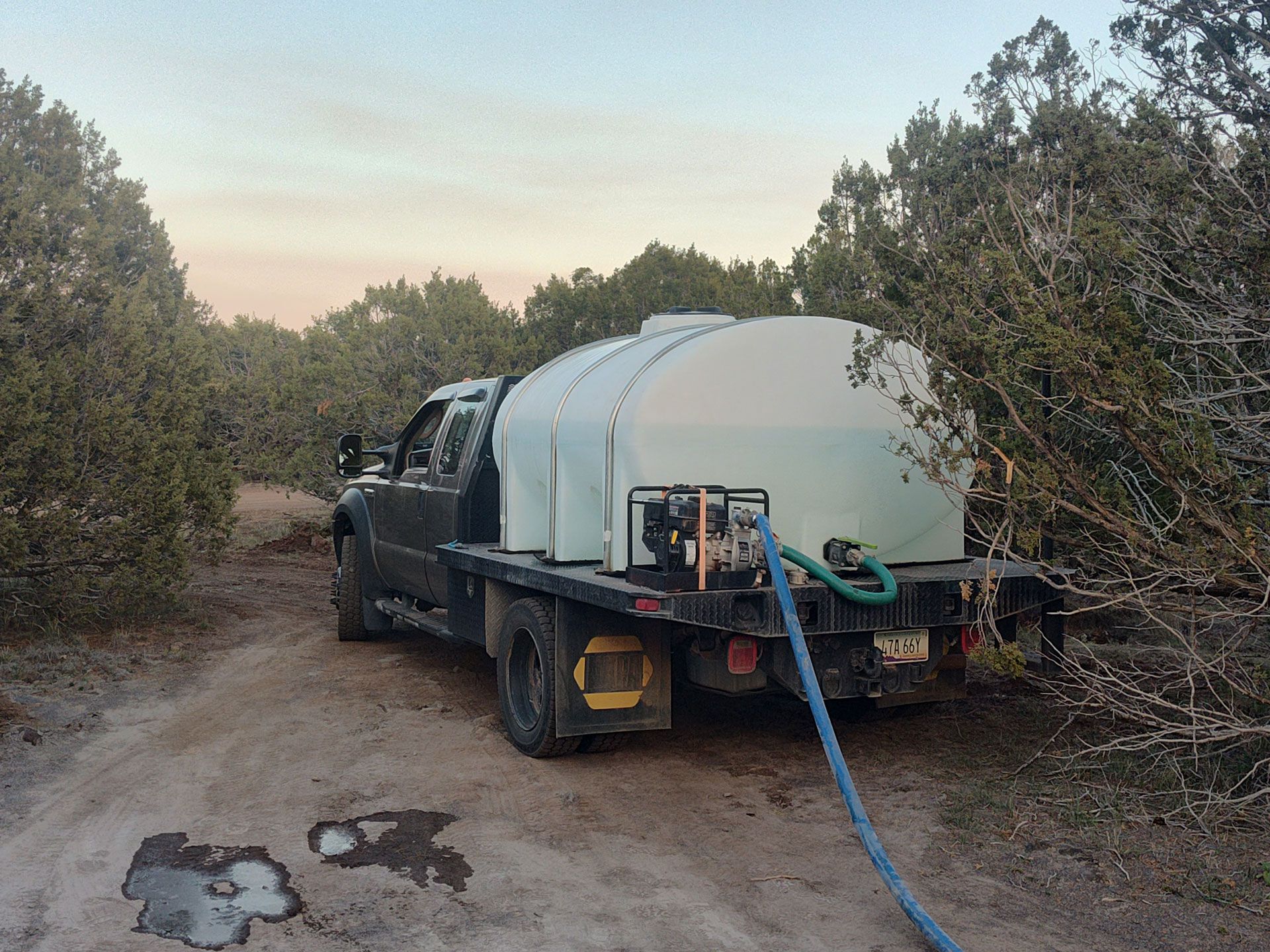 Truck-mounted water tank on a dirt road in a wooded area at dusk