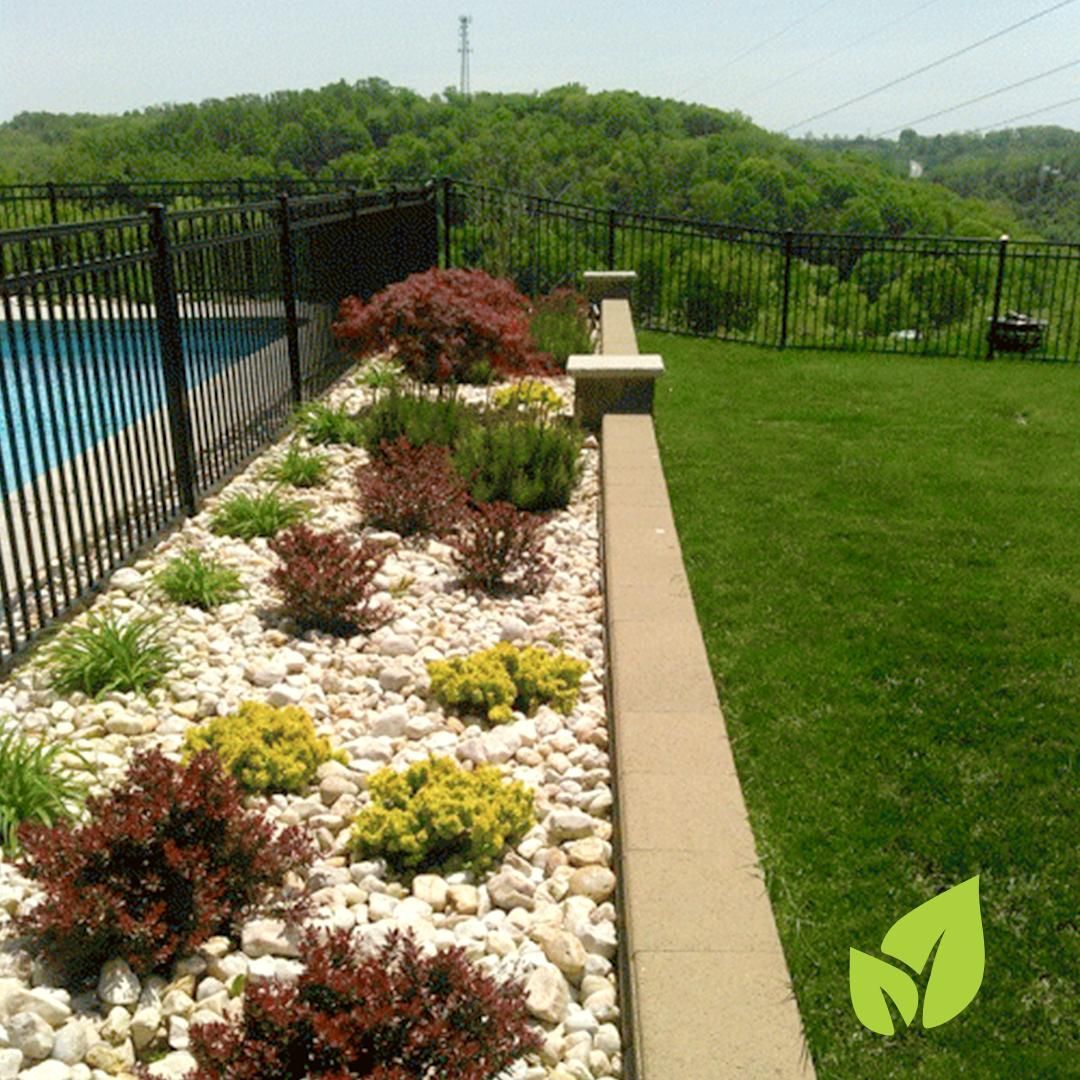 A garden bed with colorful shrubs and white stones beside a pool and lawn with a hillside in the background.
