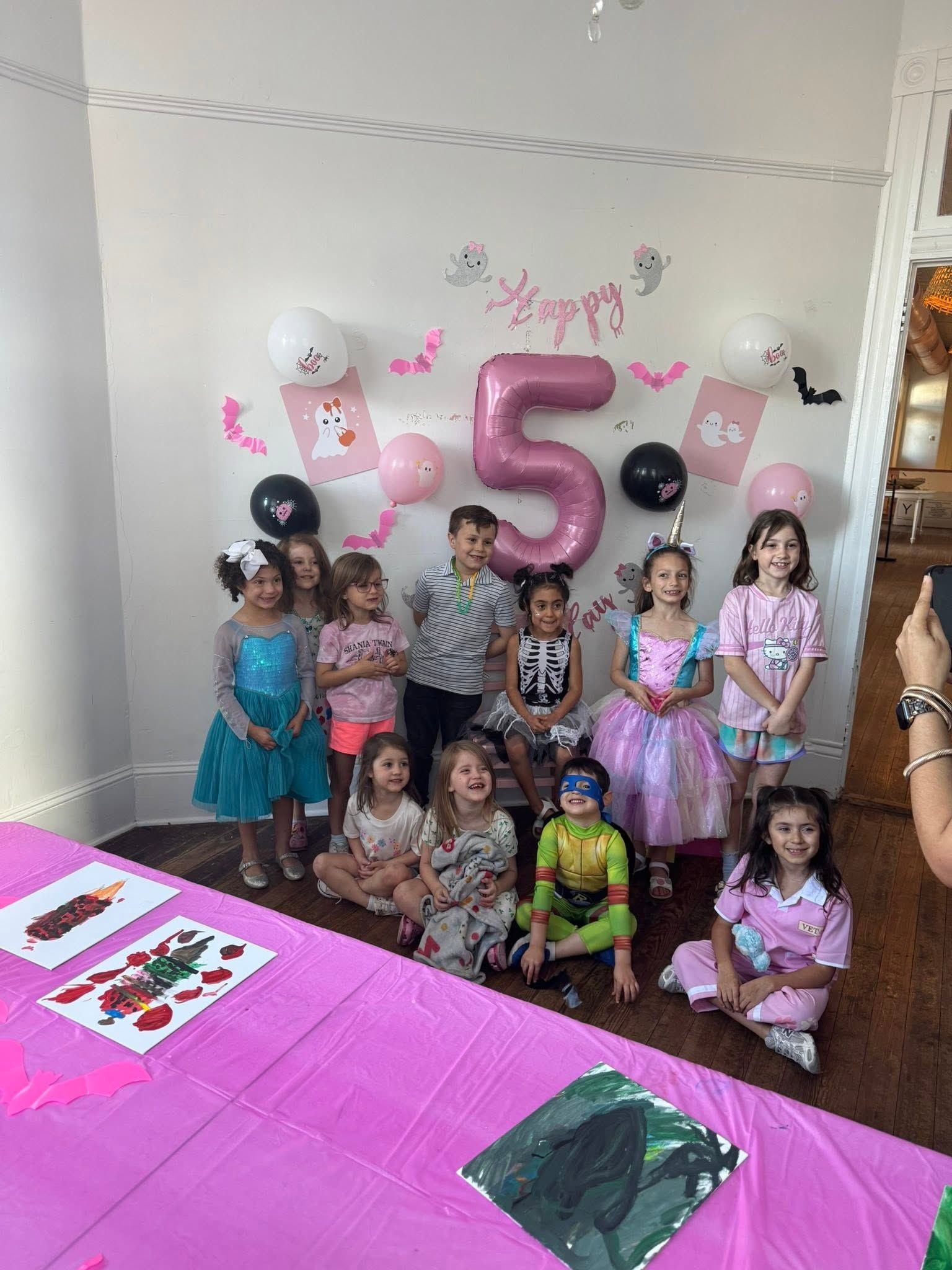 Birthday party group photo with children in colorful outfits standing before a pink “5” balloon display