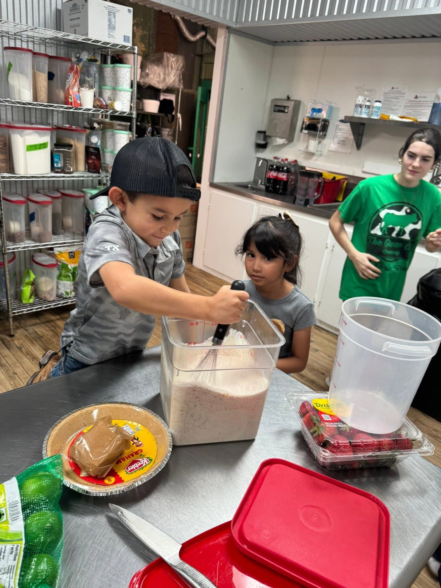 Two children mixing ingredients in a kitchen, with bowls, containers, and supplies on a counter.