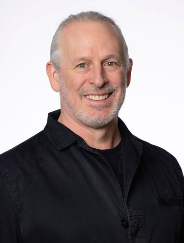 Man with graying hair and beard smiles, wearing a black button-down shirt, against a white background.