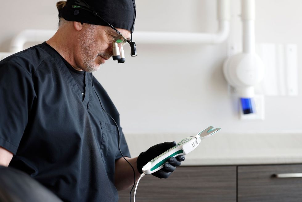 Dentist in black scrubs, wearing a headlamp and holding a dental tool, examines it in a modern office.