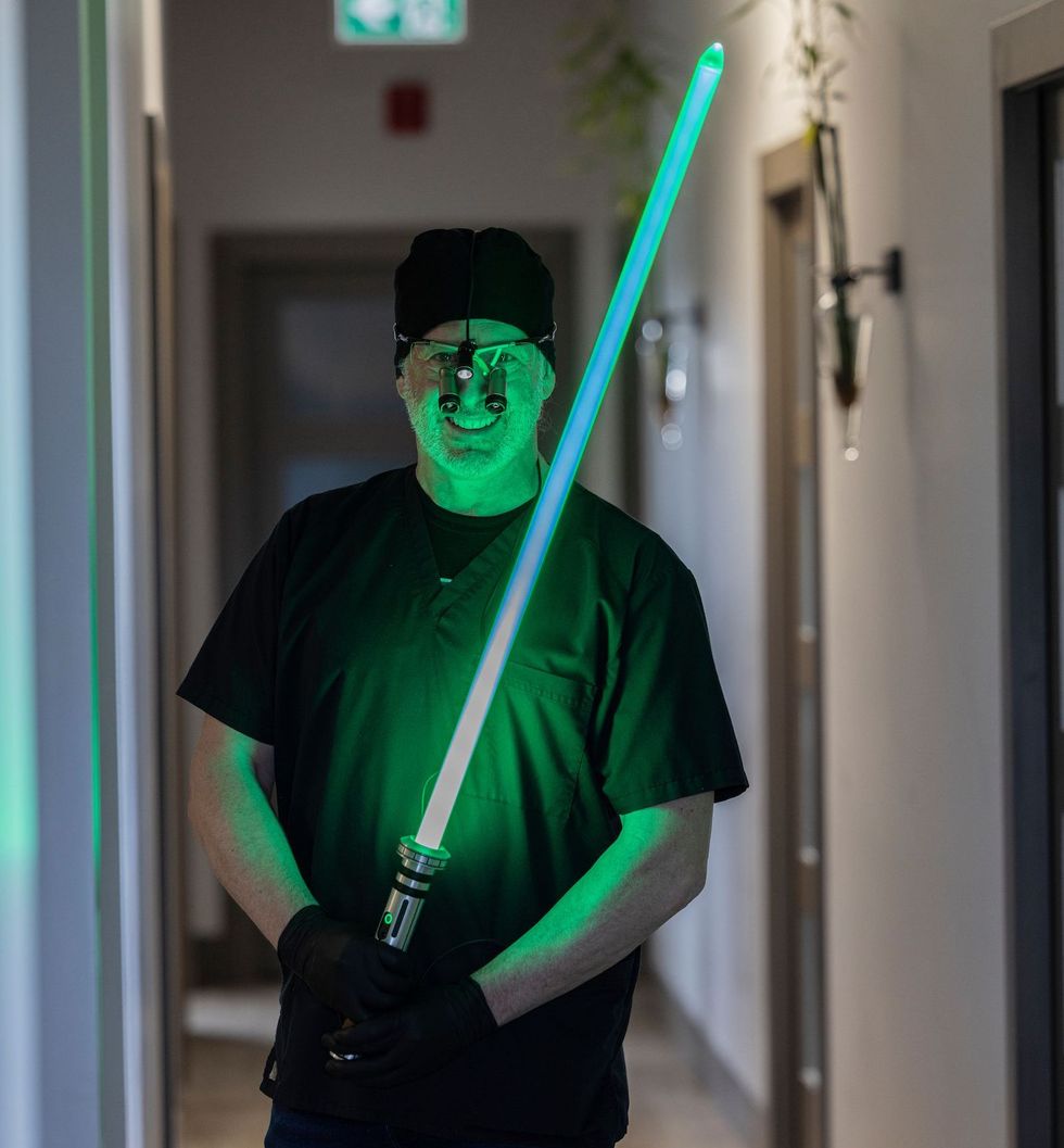 Man in scrubs holds a green lightsaber in a hallway, glowing green.