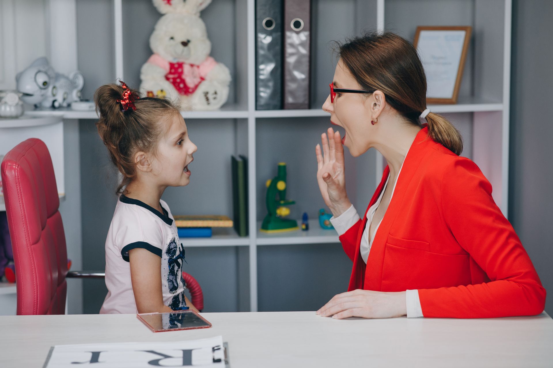 Woman in red jacket guides a girl's speech therapy; both mouth open, in a room with toys and bookshelves.