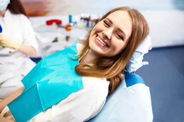 Woman in dental chair smiling, blue bib, dentist in background.