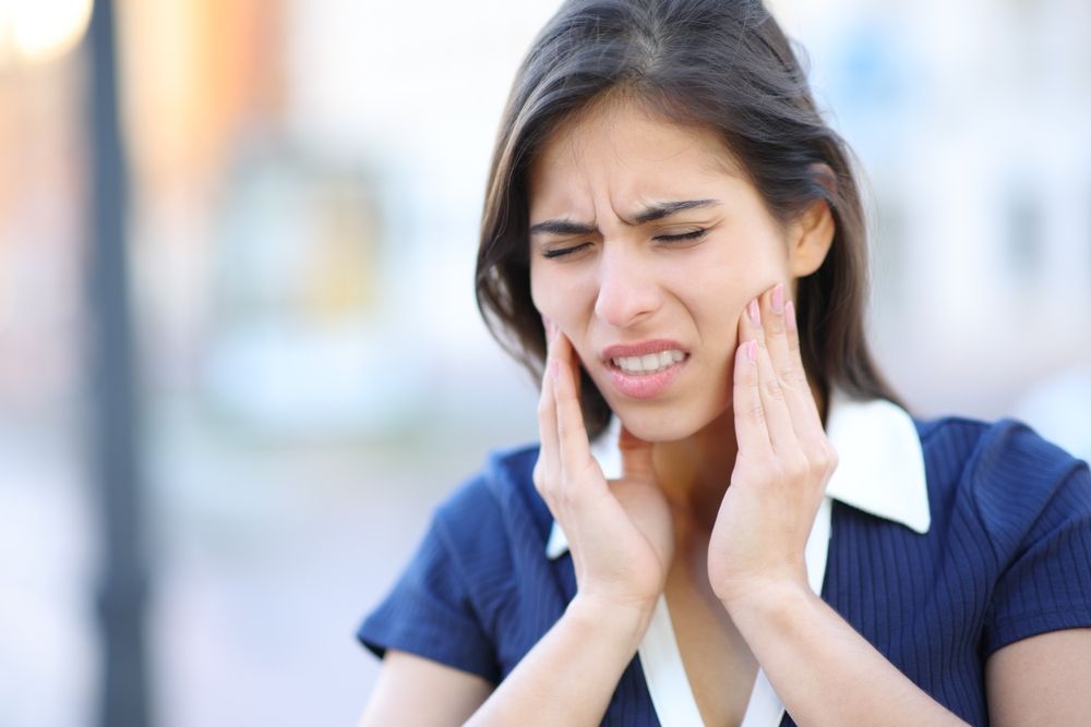 Woman in blue top with pained expression, hands on cheeks, outdoors, possibly in pain.