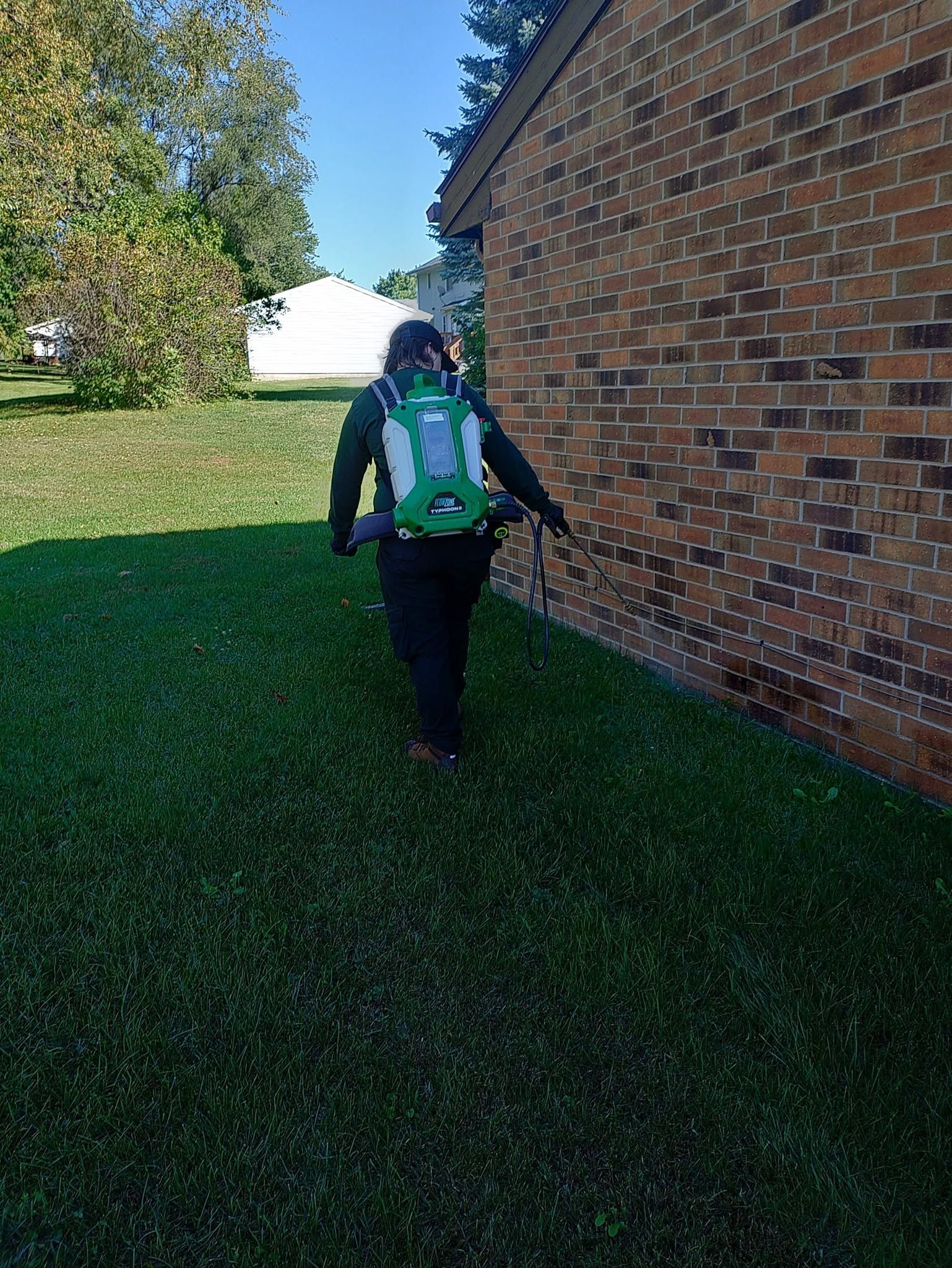 Person in a green jacket walking beside a brick building on a grassy lawn.
