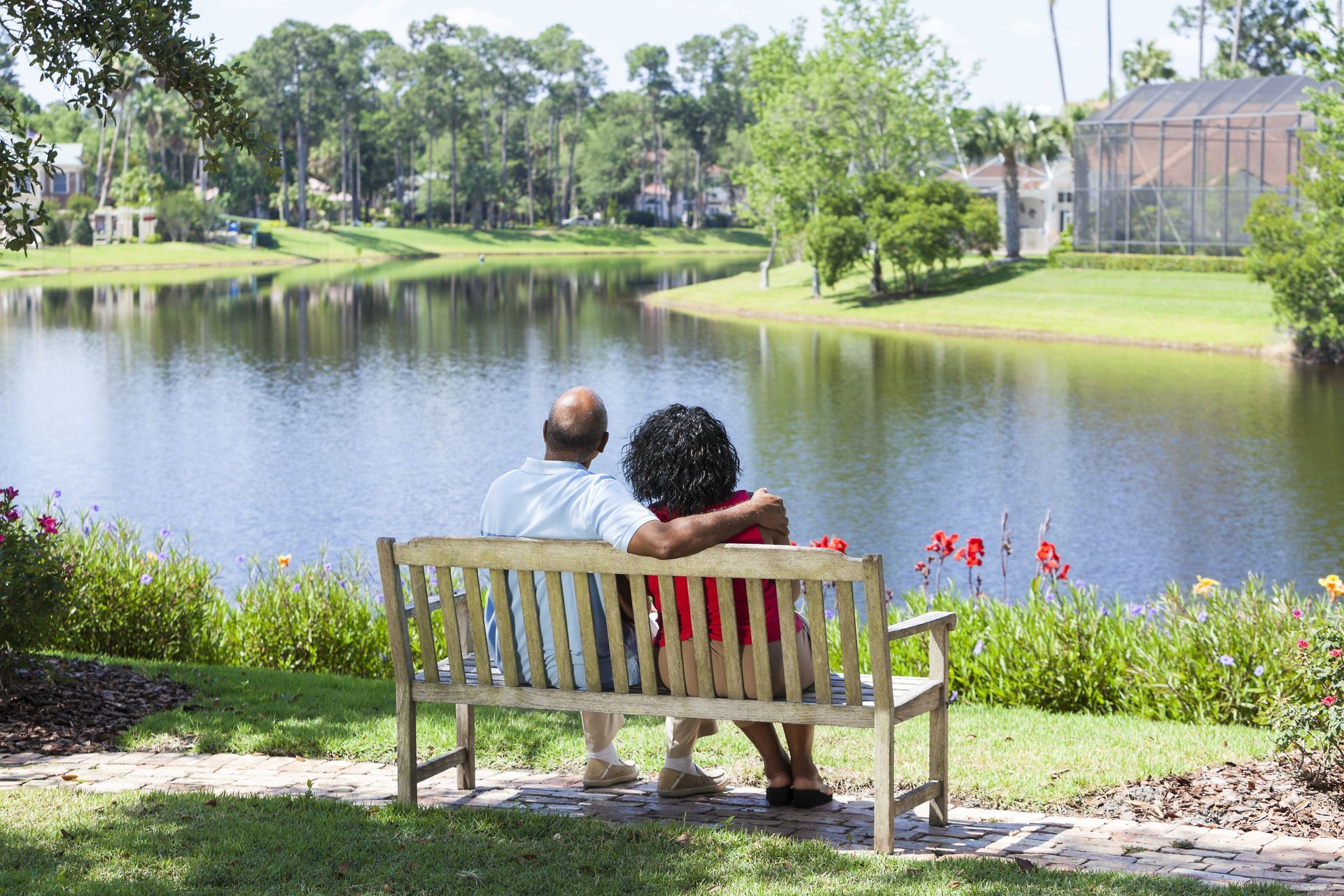 Couple sitting on a bench by a pond in a sunny park, with trees and flowers in the background