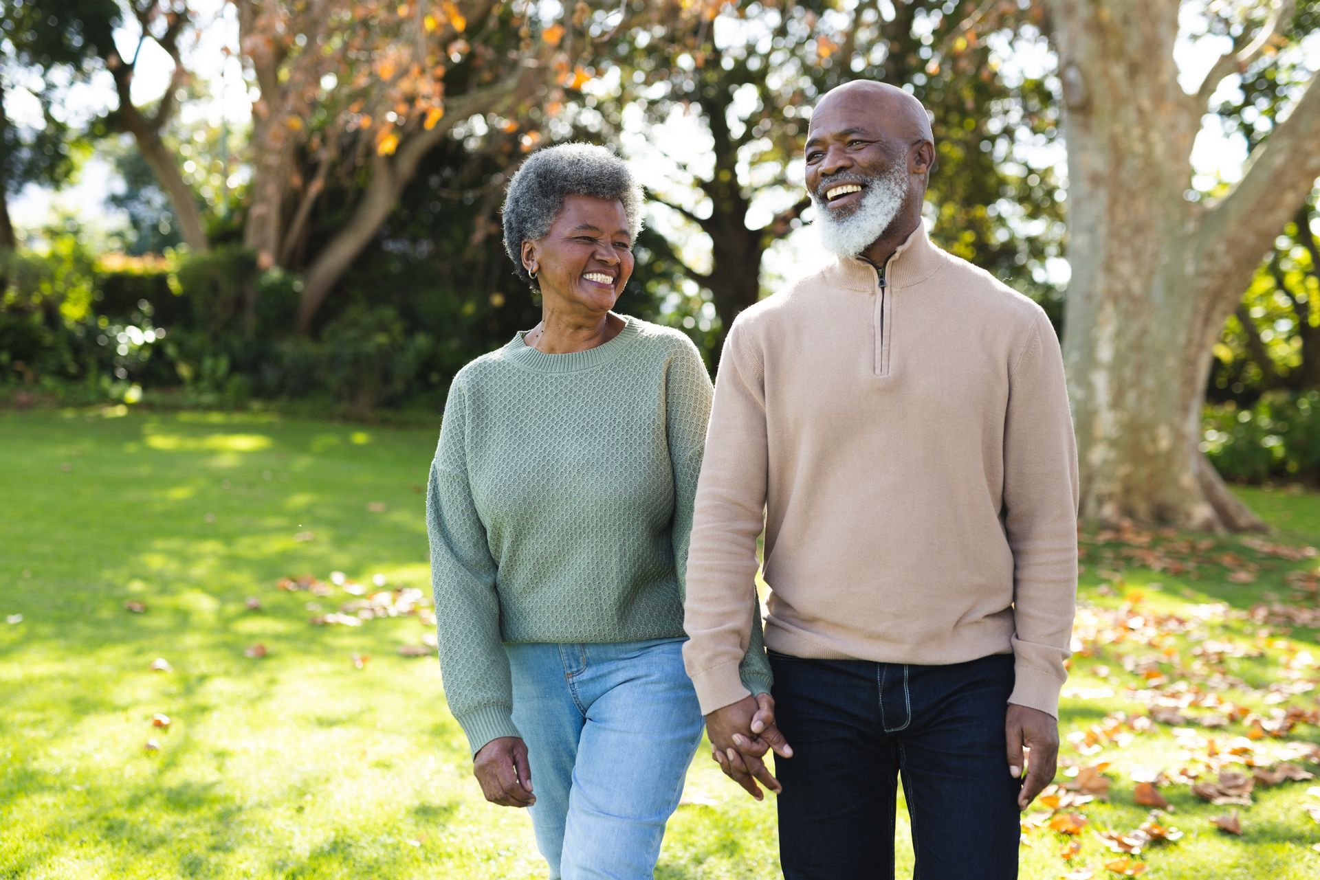 Two smiling people walking together on a sunlit park path, with trees and grass in the background.