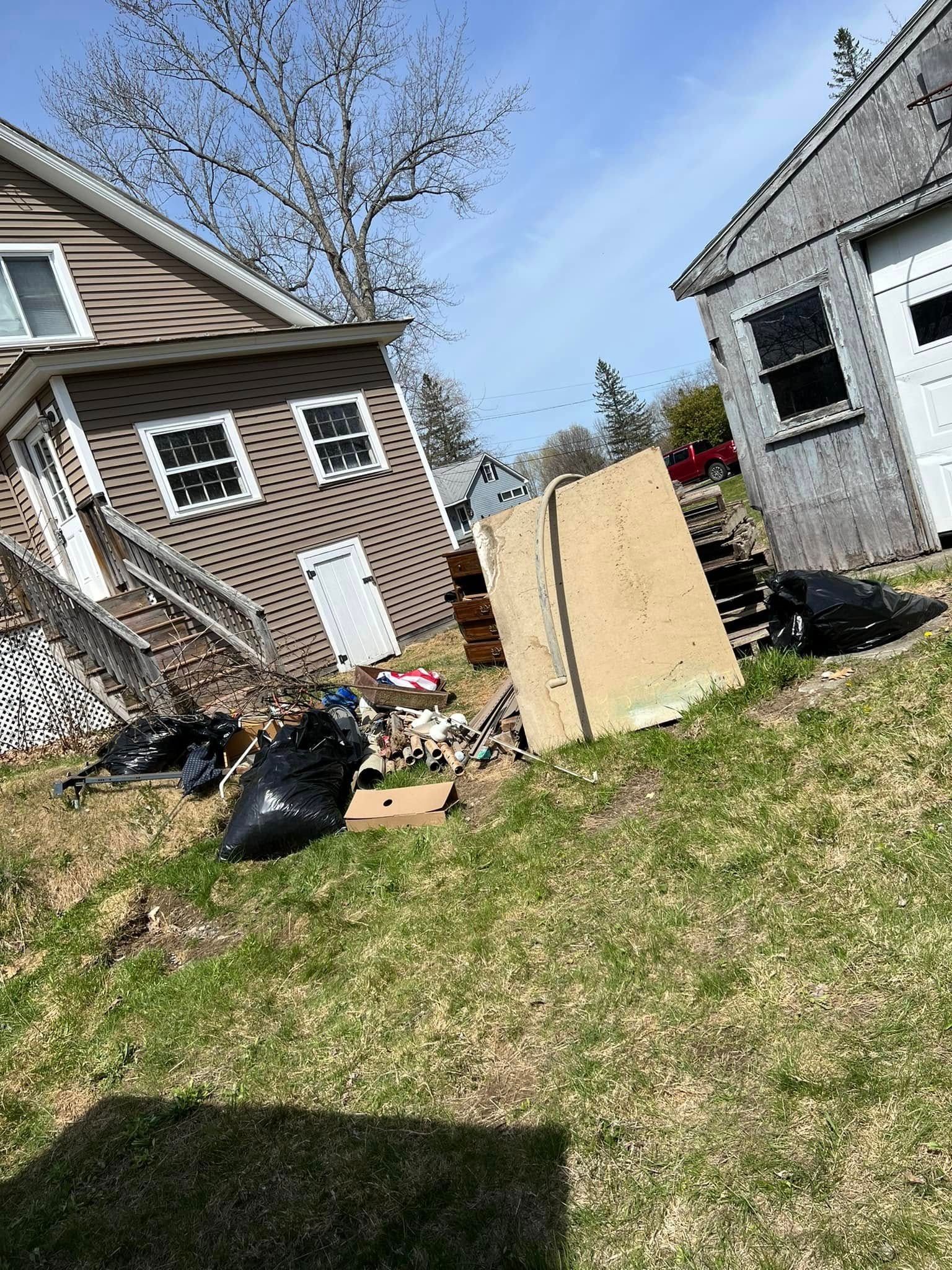 Tilted backyard scene with debris, black trash bags, a wooden panel, and two houses under a blue sky
