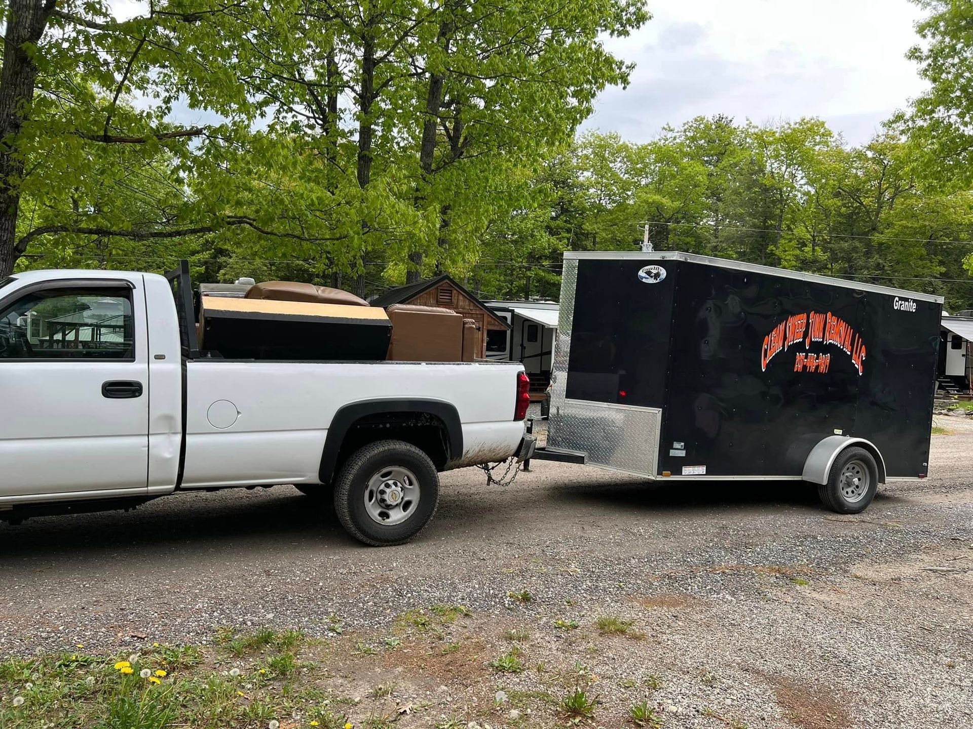 White pickup truck towing a black enclosed trailer on a gravel road by trees.