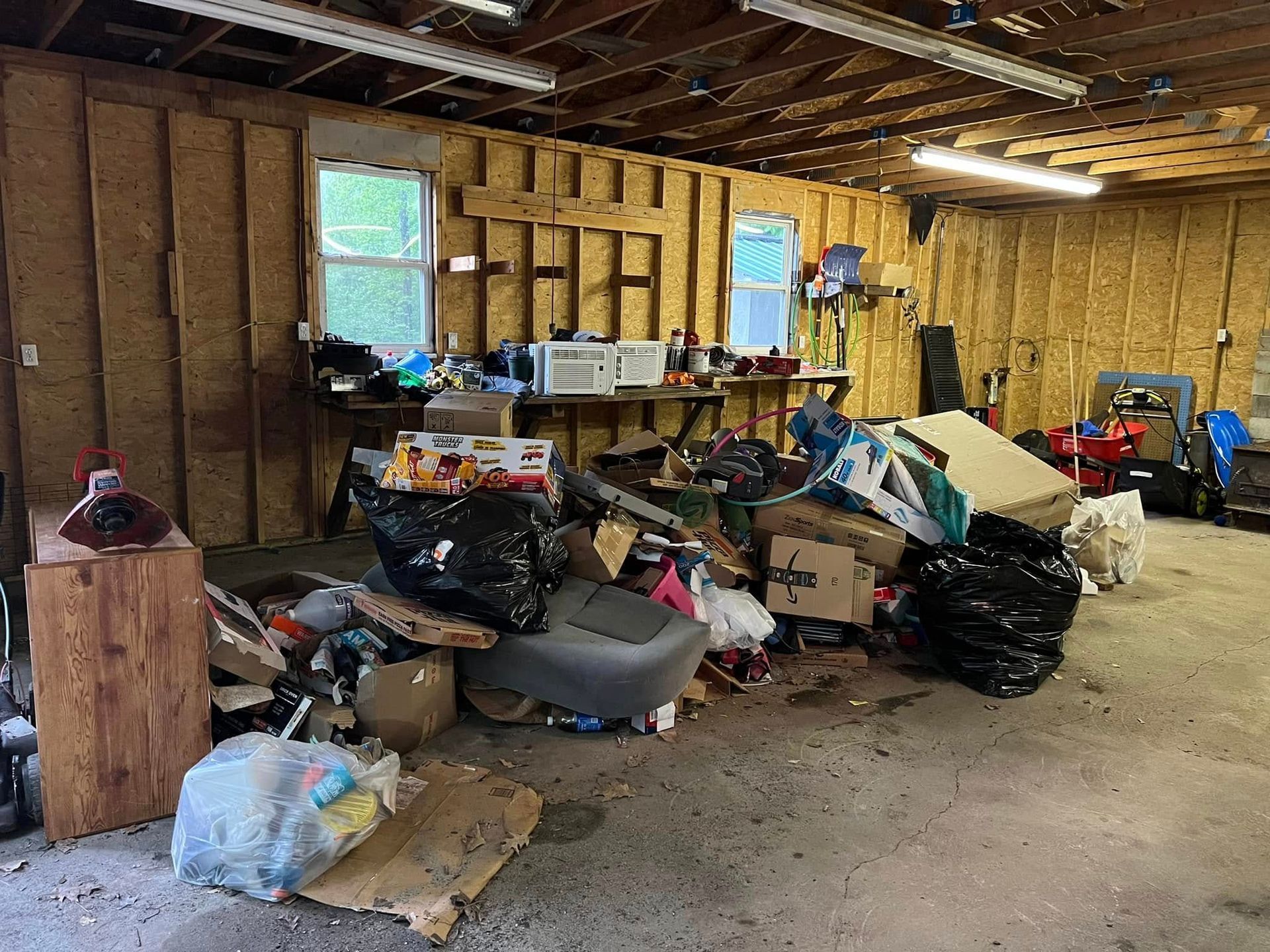 Cluttered basement with piles of trash, bags, and household debris along unfinished wooden walls.