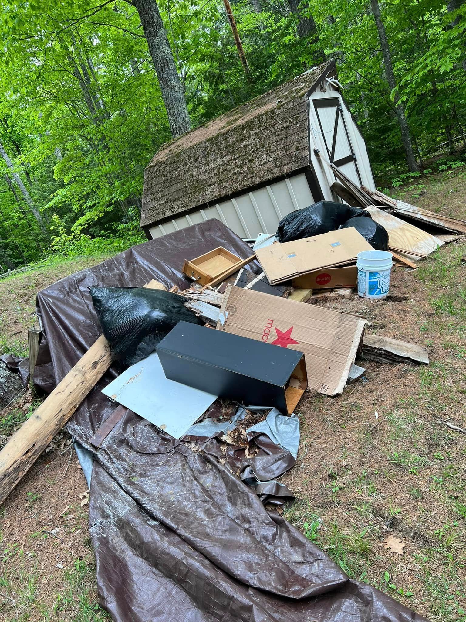 Bulky yard debris and broken furniture piled beside a small shed in a wooded area