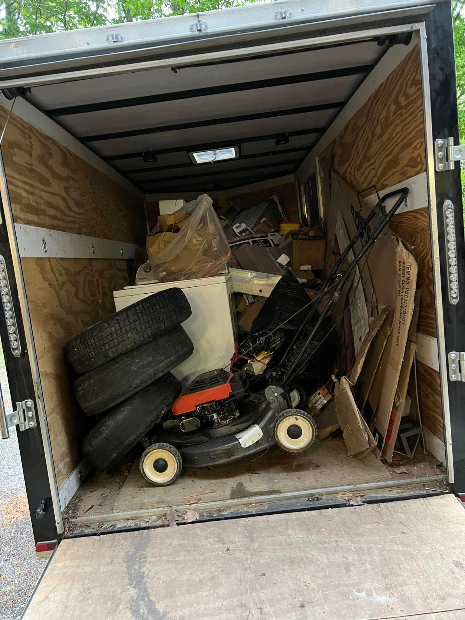 Open trailer packed with debris, tires, and a black lawn mower inside wood-paneled walls.