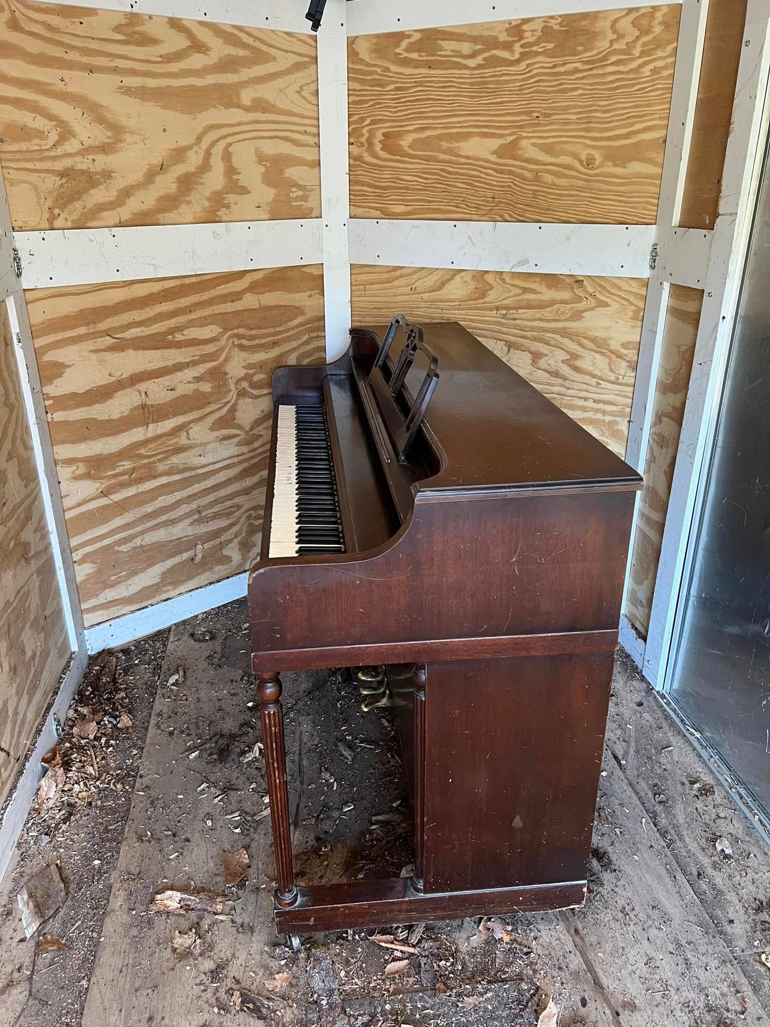Worn upright piano inside a plywood storage crate, with exposed keys and dusty floor.