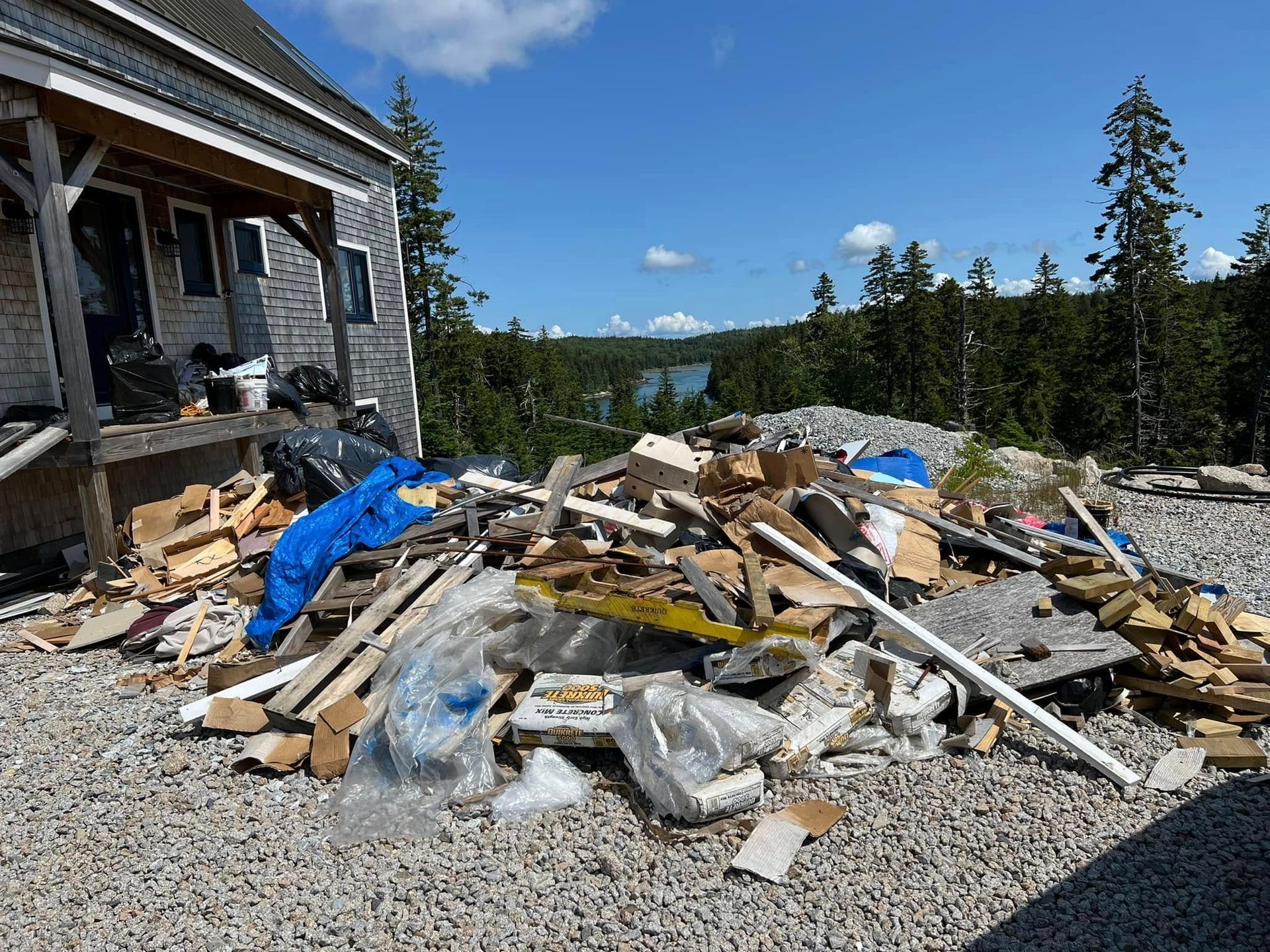 Pile of construction debris beside a stone house under a blue sky and trees