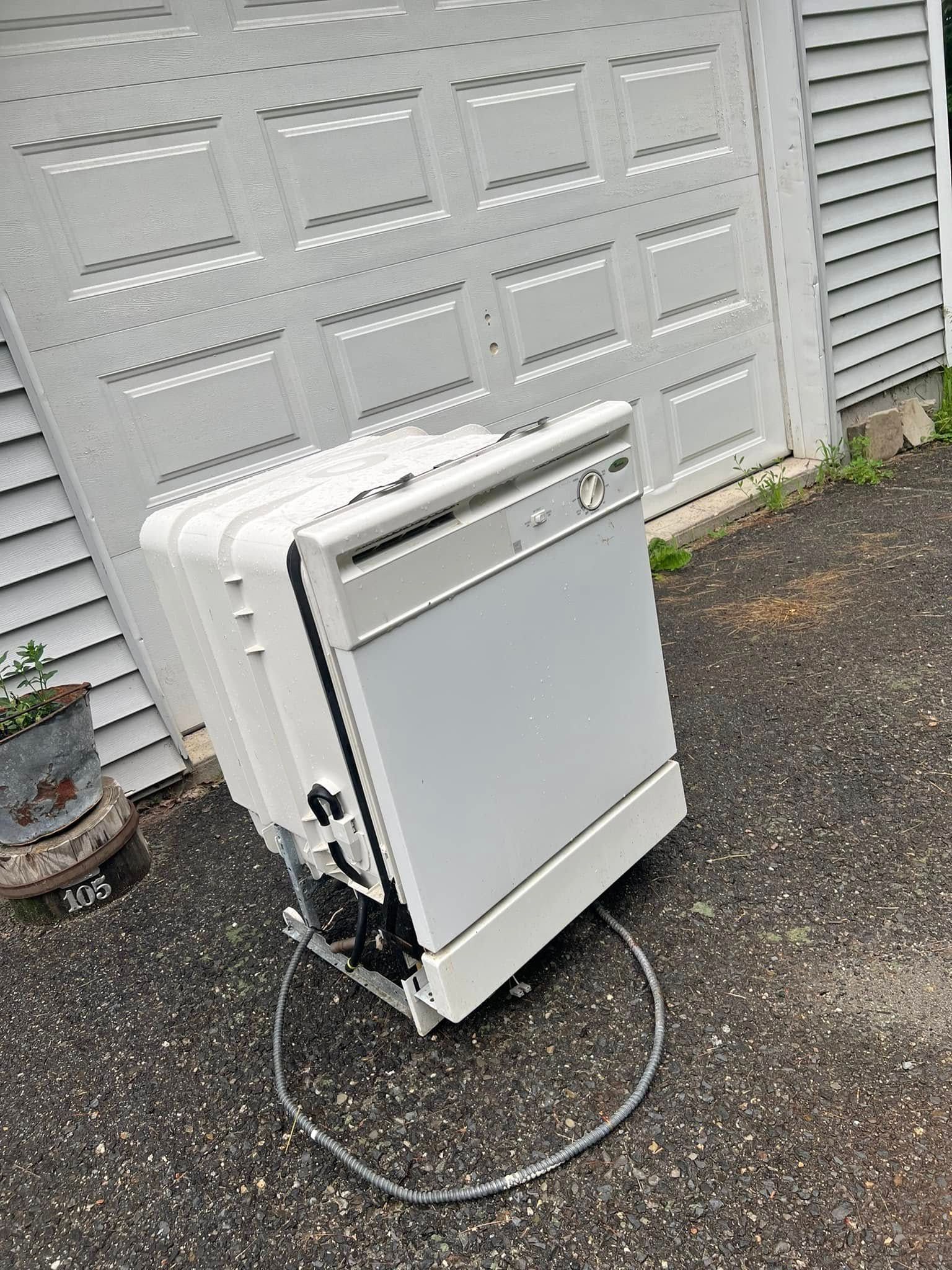 Portable dishwasher unit on wet gravel beside a house, with hose attached and a white siding wall behind.