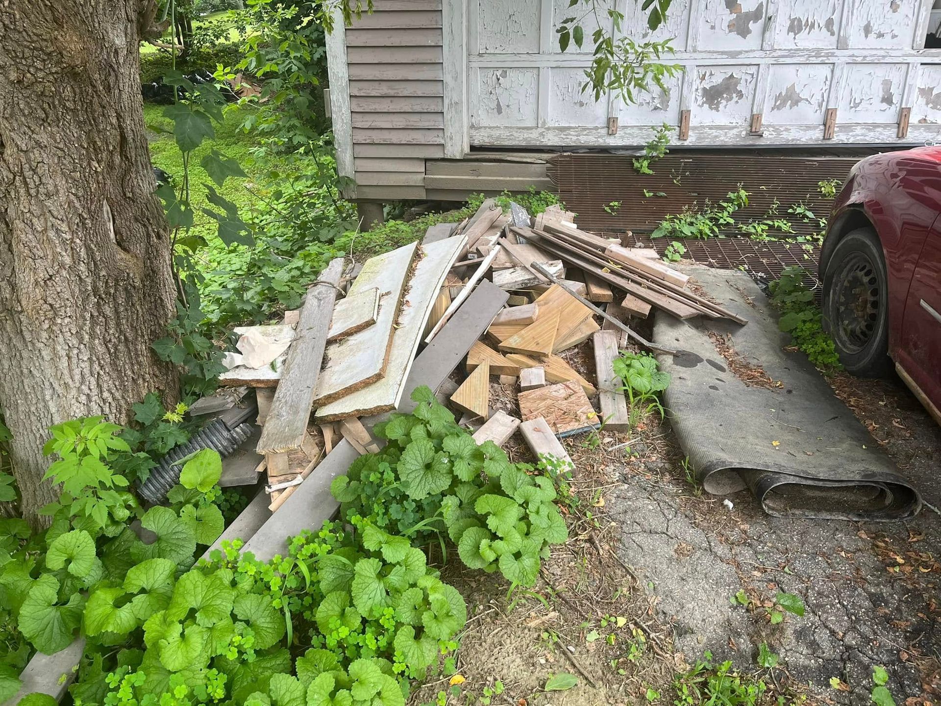 Pile of broken boards and debris beside a house, with a tree and red car nearby.