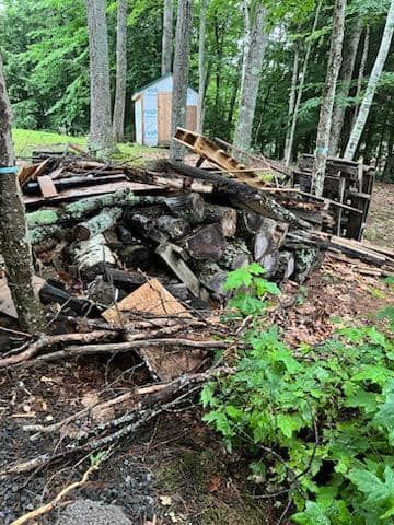 Woodpile and stacked scrap wood beside a small shed in a wooded backyard
