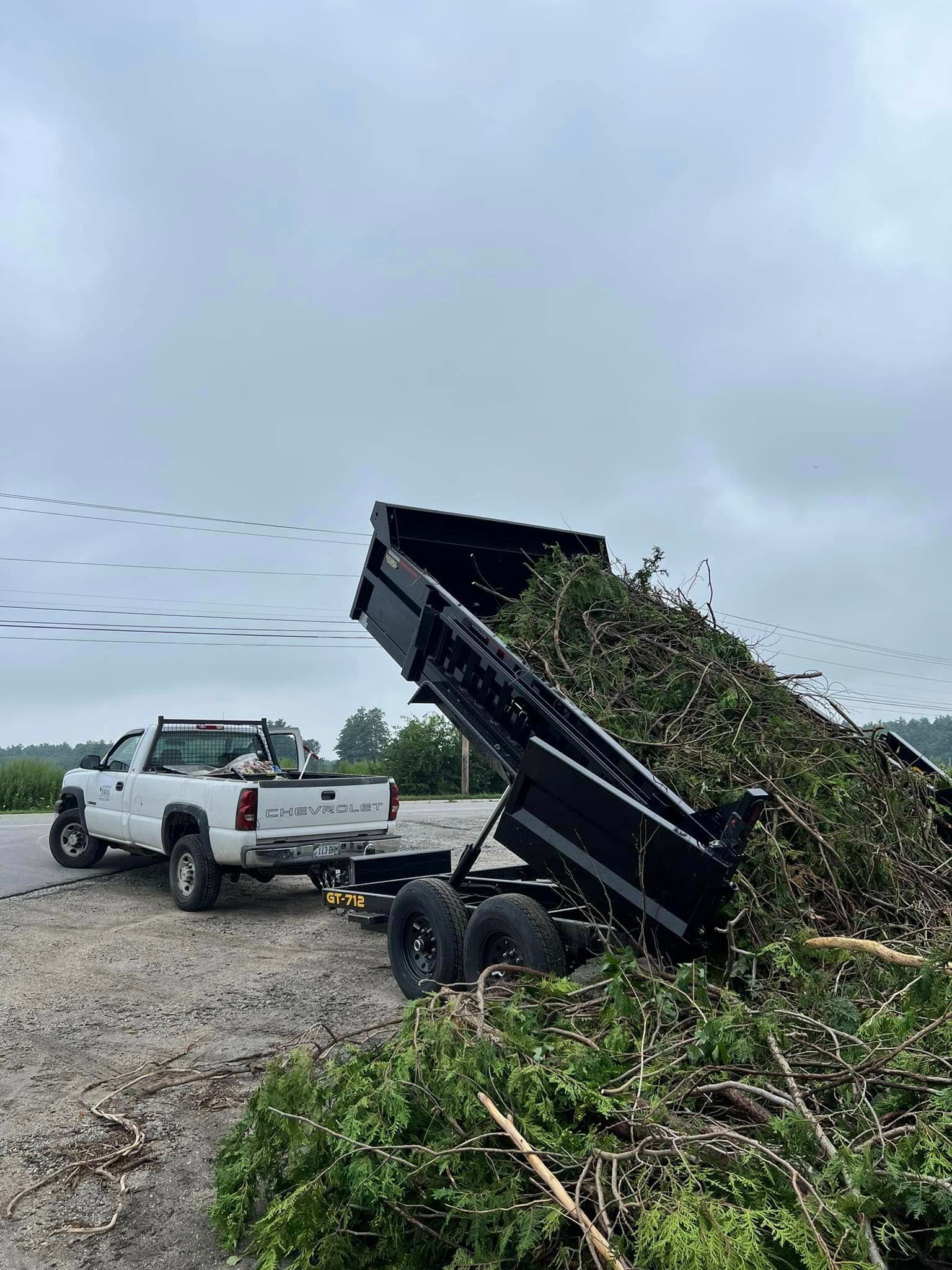 Dump truck unloading green yard waste beside a white pickup on a gravel road under cloudy sky