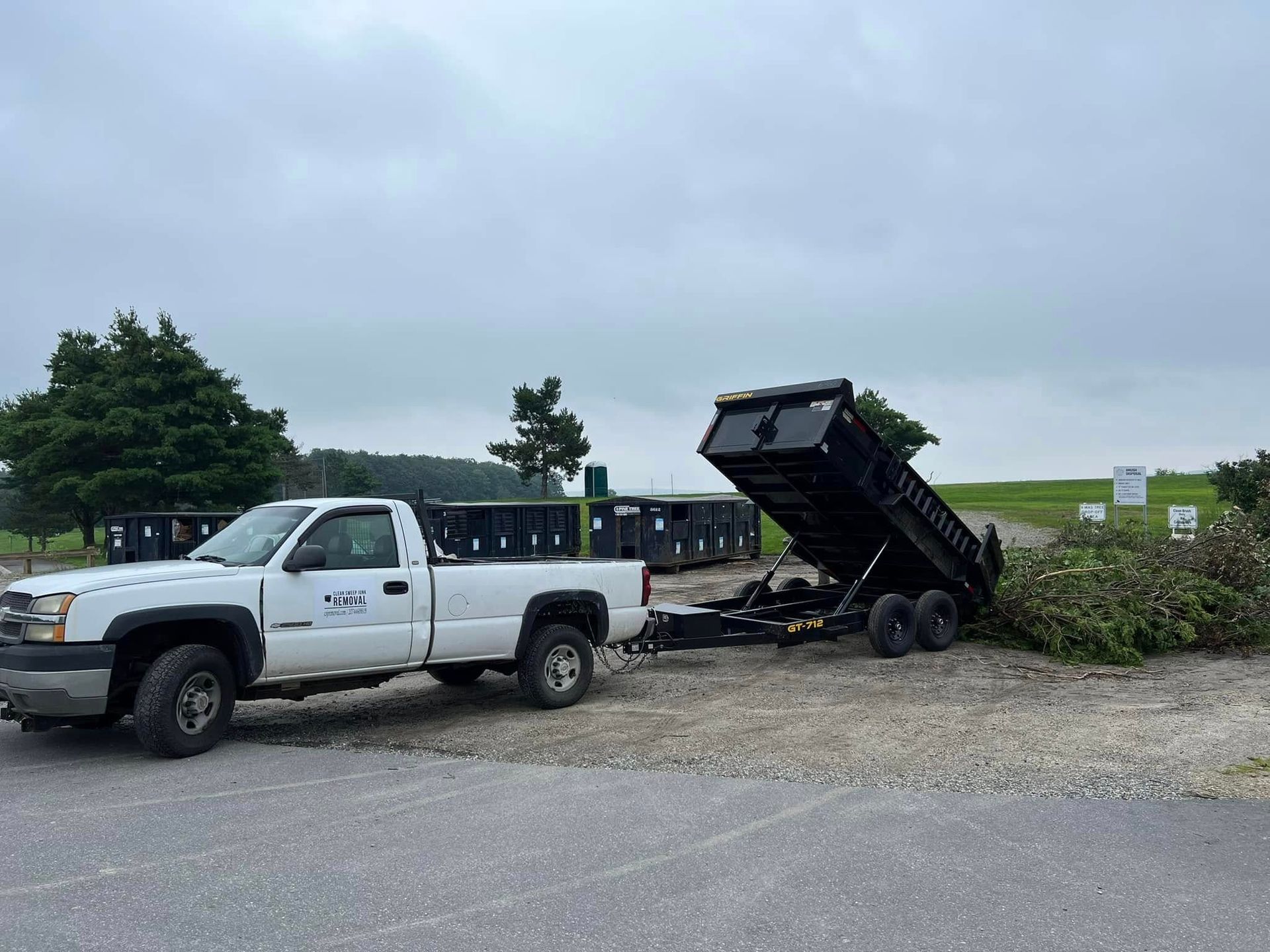 White pickup truck beside a dump trailer tipped up in a gravel lot under a cloudy sky