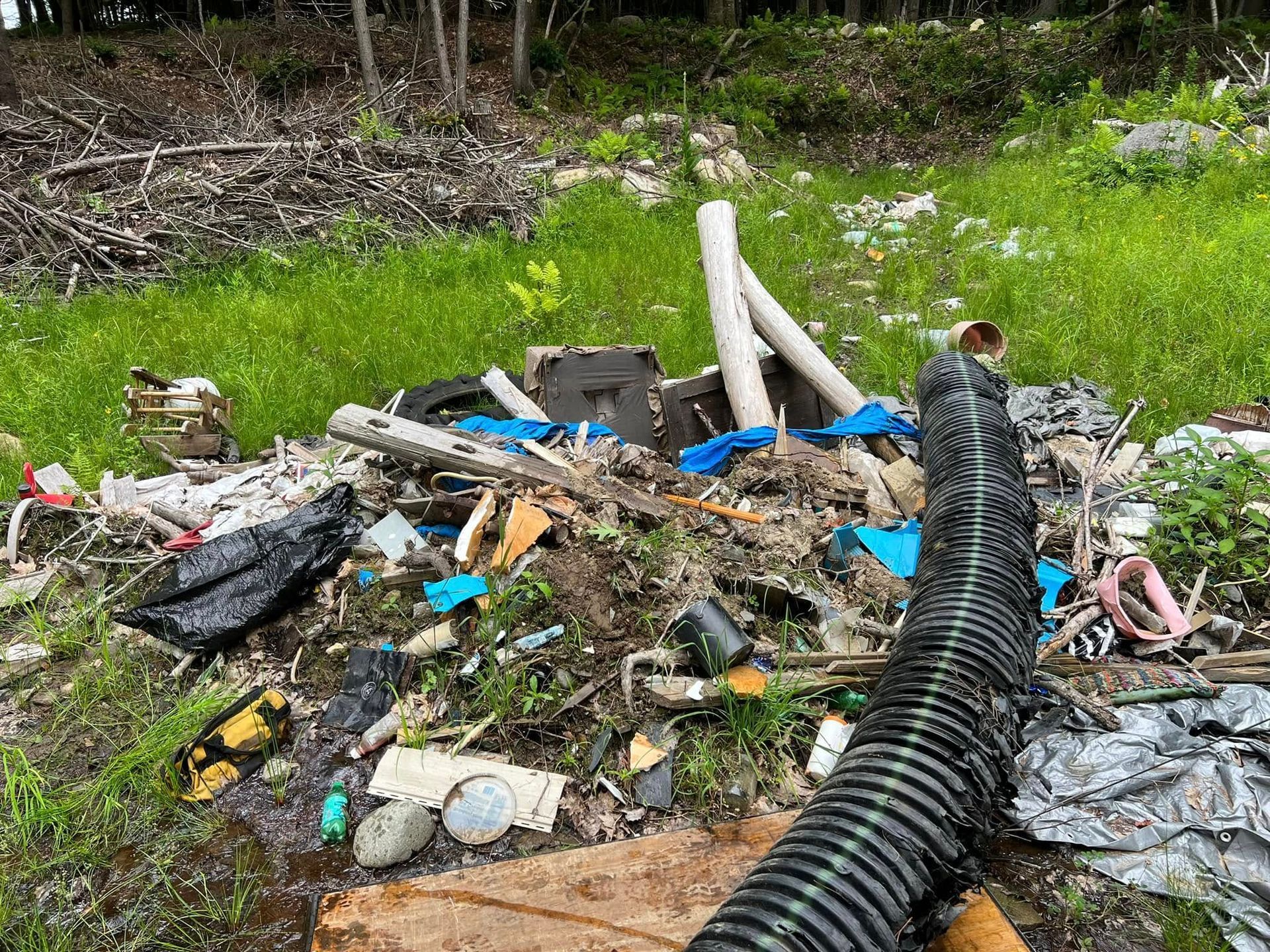 Pile of mixed trash and debris scattered in a grassy outdoor area with pipes and plastic bags.