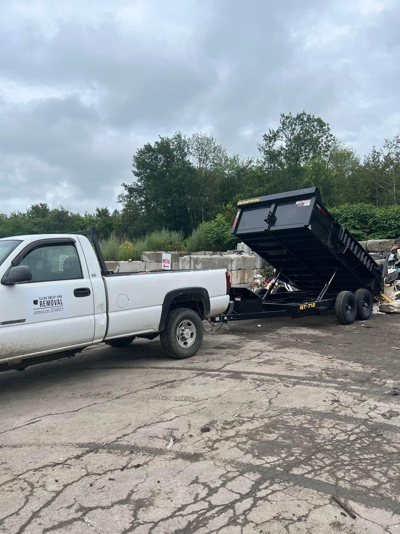 White pickup truck hauling a black dump trailer on cracked pavement under cloudy skies