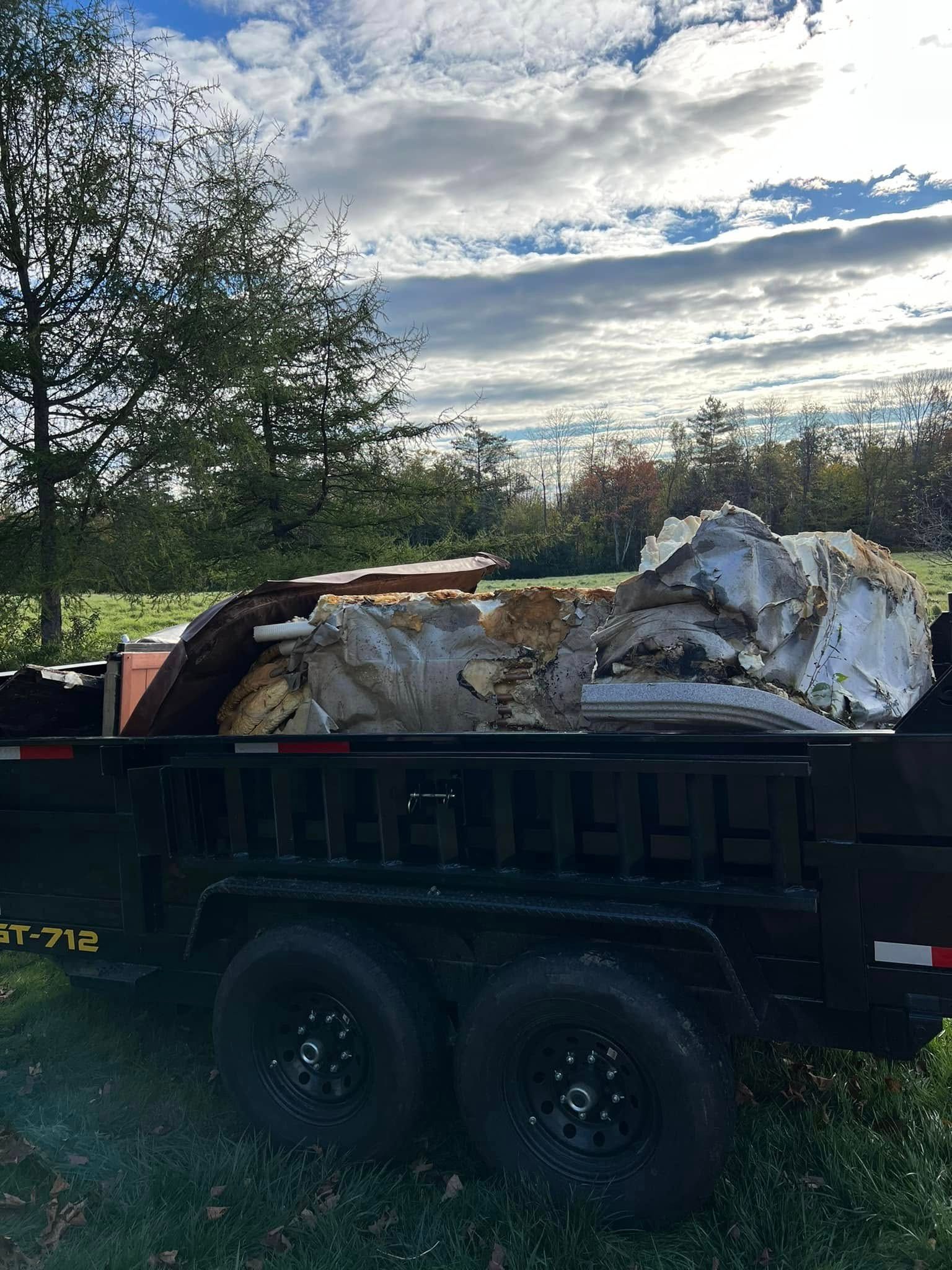 A black dump trailer loaded with debris sits in a grassy field under a cloudy sky.