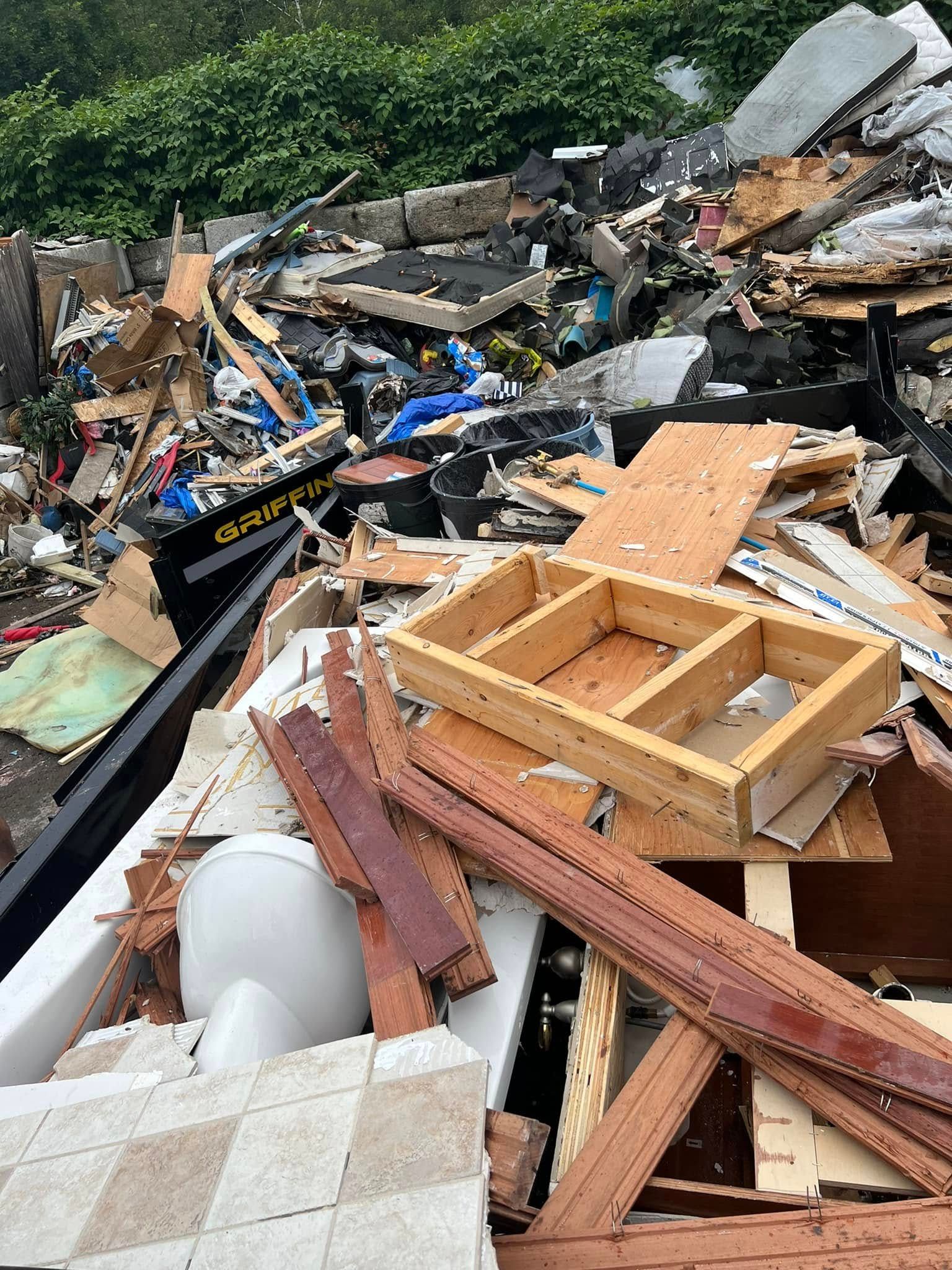 Pile of construction debris and broken wood scattered outdoors beside a green hedge.