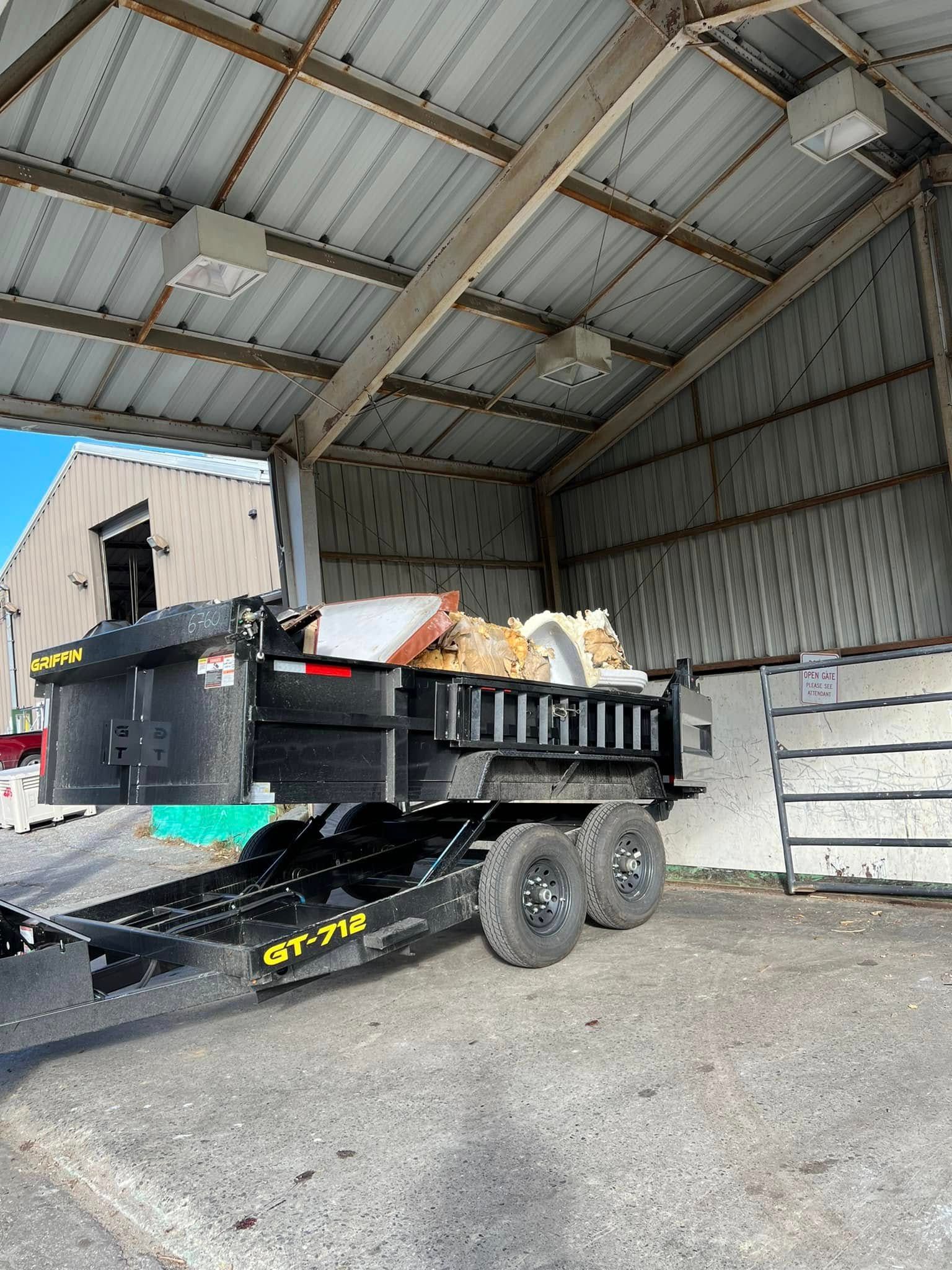 Black dump trailer loaded with debris inside a large metal shed, with stacked concrete blocks nearby.