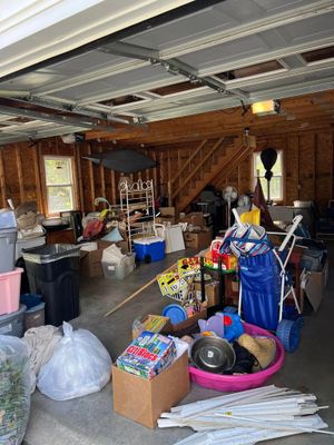 Cluttered garage with storage bins, boxes, tools, and a staircase under an open ceiling