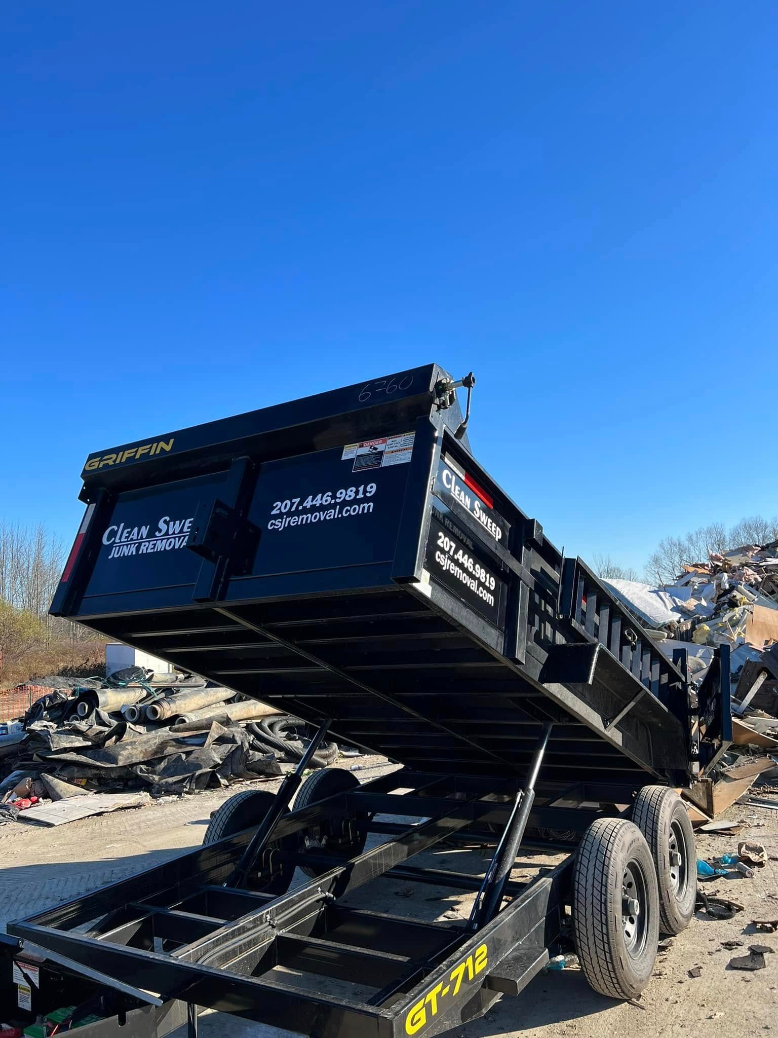 Dump trailer tilted upward in a debris-strewn lot under a clear blue sky.
