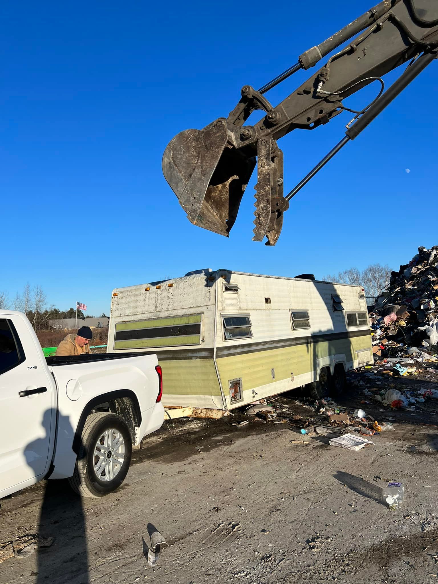Excavator bucket lifting debris beside a white pickup and camper trailer at a scrapyard under a blue sky