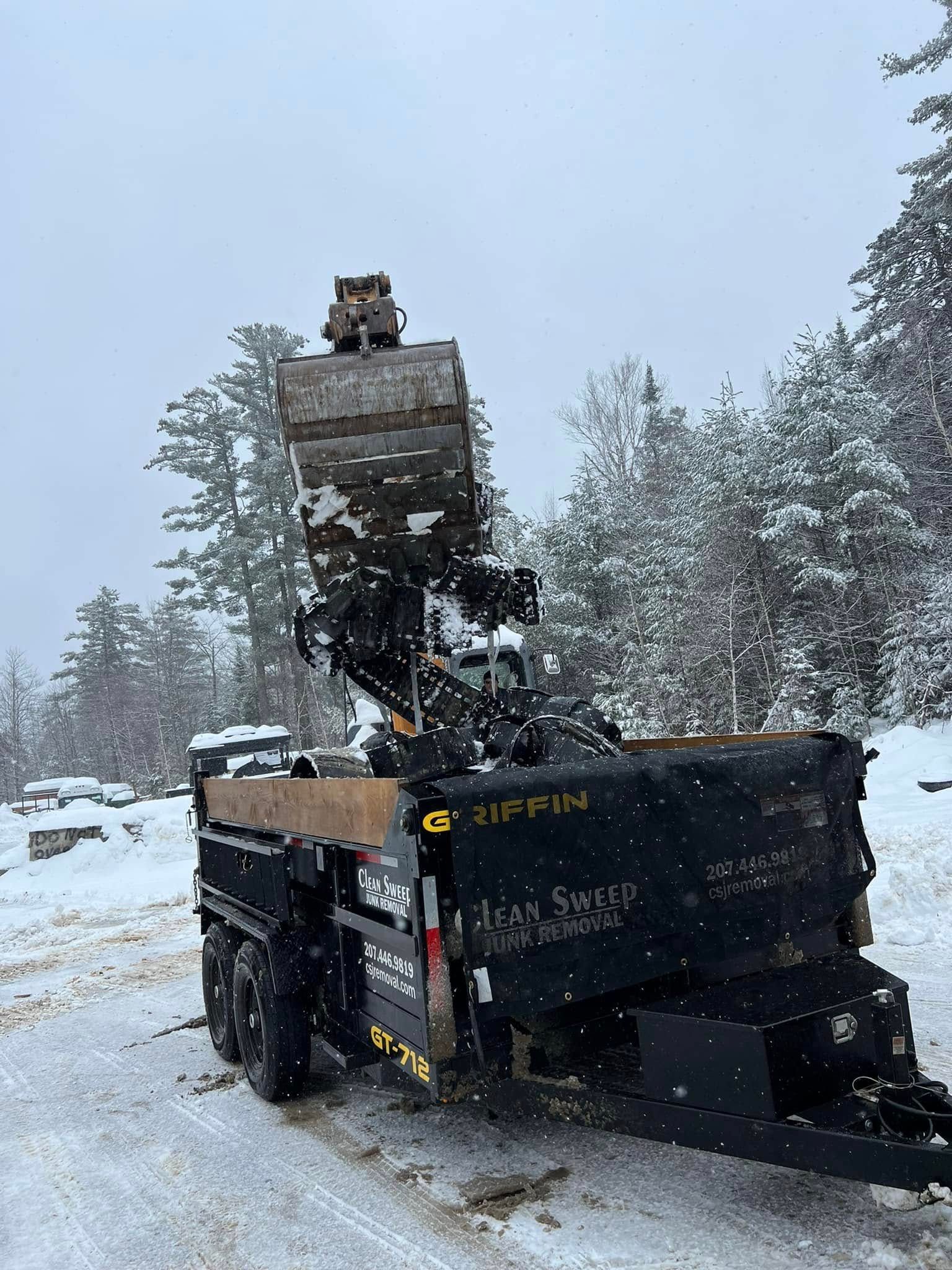 A snow-covered dump truck unloading material beside a snowy road, with a raised bed and loader overhead
