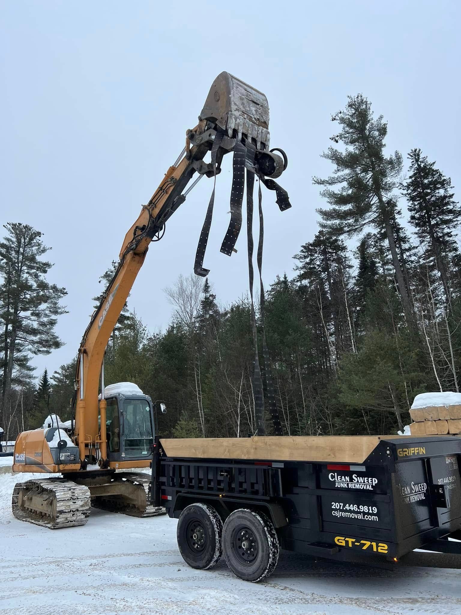 Excavator lifting a black truck chassis in a snowy forest work site