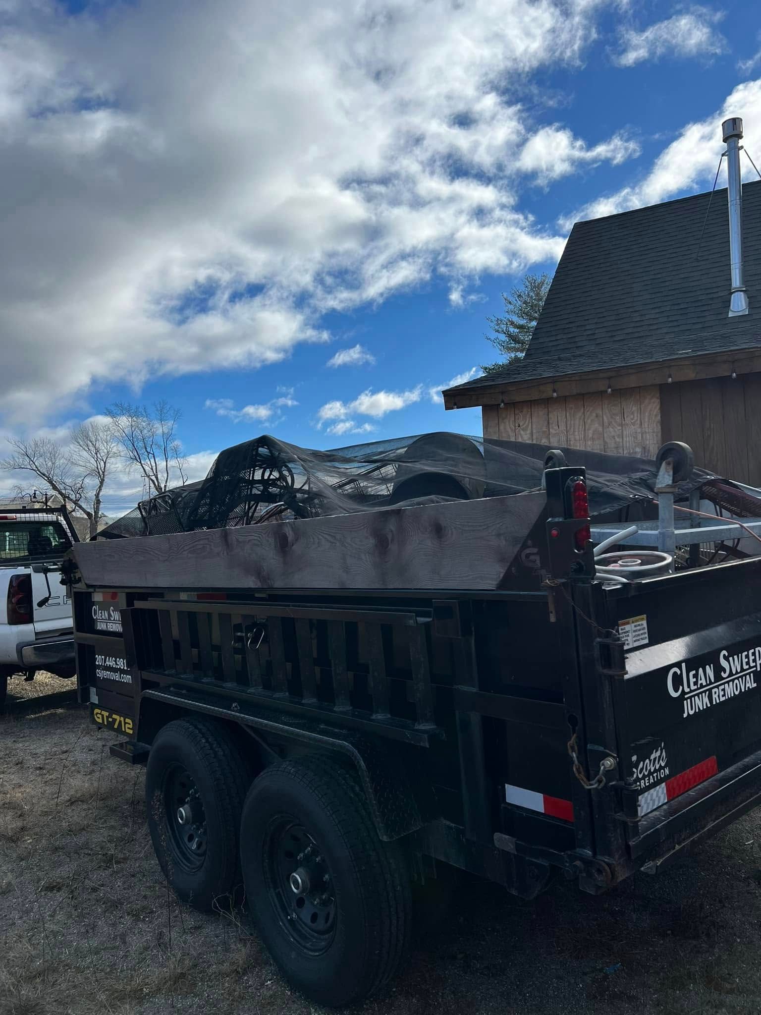 Black dump trailer filled with debris parked beside a house under a cloudy blue sky